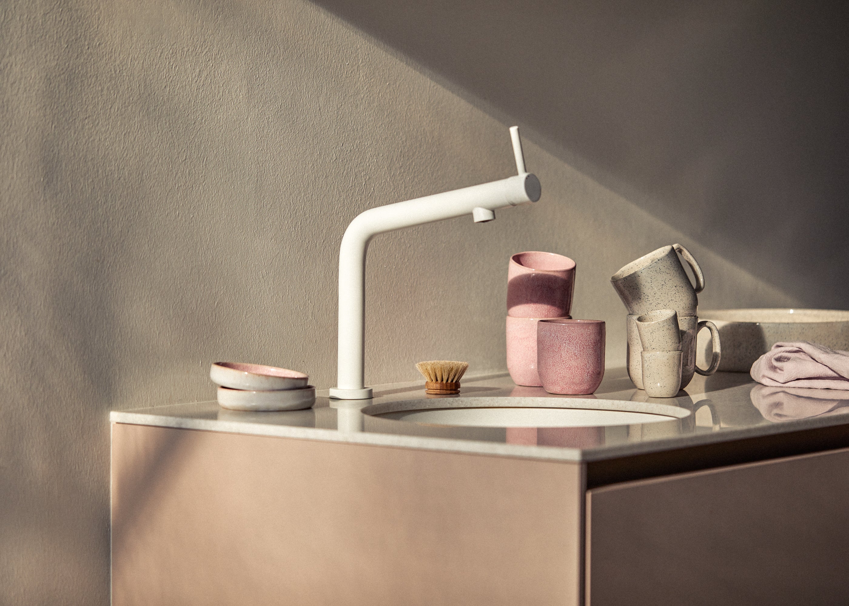 Modernes Küchenspülbecken aus hellem Stein mit weißer Design-Armatur, daneben rosa und cremegrau gesprenkeltes Keramikgeschirr (Becher mit und ohne Henkel sowie kleine Teller), eine Holzspülbürste und ein gefaltetes lavendelfarbenes Geschirrtuch im warmen Sonnenlicht.