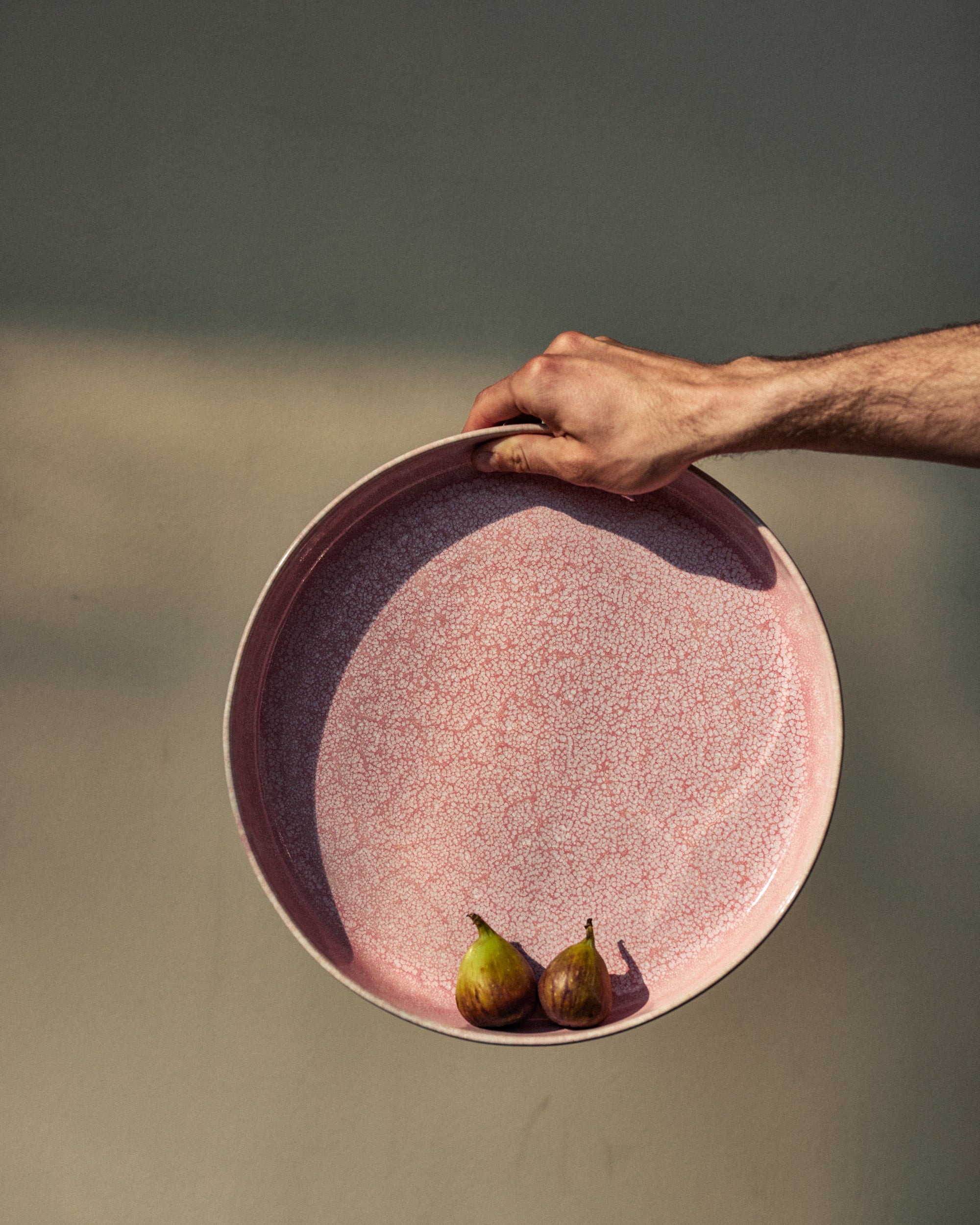 Hand hält eine große runde Keramikschale mit rosa-weißer Craquelé-Glasur und dunkelviolettem Rand, darin zwei frische grüne Feigen. Vor unscharfem grau-grünem Hintergrund im Sonnenlicht.