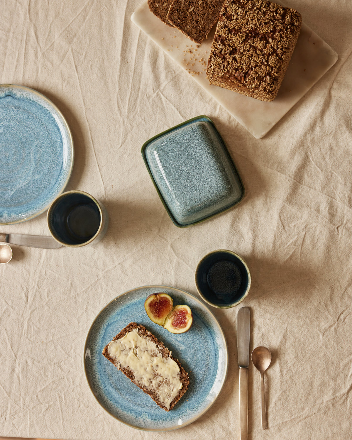 Frühstückstisch von oben auf beigem Leinentischtuch: Ein blauer Keramikteller mit gebutterter Brotscheibe und aufgeschnittenen Feigen, daneben eine grün-blaue Keramik-Butterdose, zwei dunkle Espressobecher und silbernes Besteck. Im Hintergrund aufgeschnittenes Körnerbrot auf einem Marmorschneidebrett.