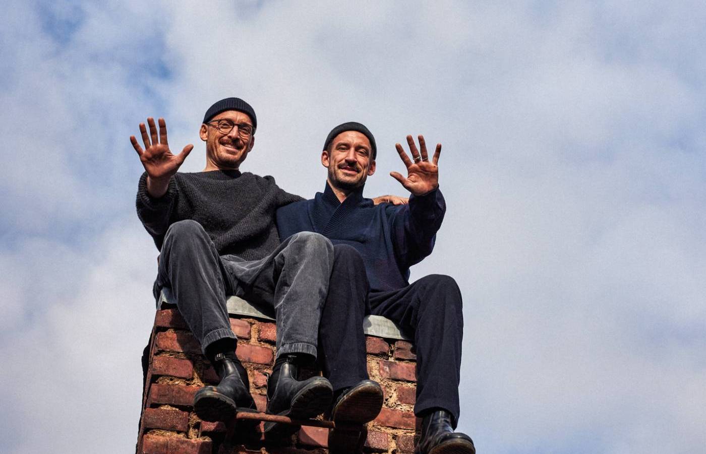 Zwei Männer sitzen lachend und winkend auf einem Backsteinschornstein vor bewölktem Himmel