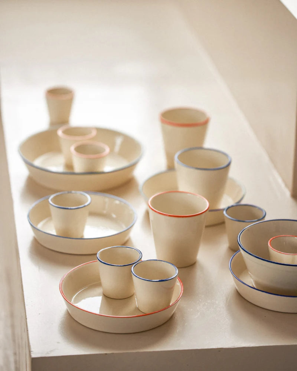 Draufsicht auf eine Kollektion cremefarbener Keramikstücke auf einem weißen Regal. Die Serie umfasst runde Teller mit integrierten Kerzenhaltern, konische Becher sowie kleine Schüsseln – alle mit einem feinen Rand in abwechselnd Blau oder Korallrot akzentuiert. Das schlichte, handgefertigte Design wirkt durch das weiche Seitenlicht besonders elegant.