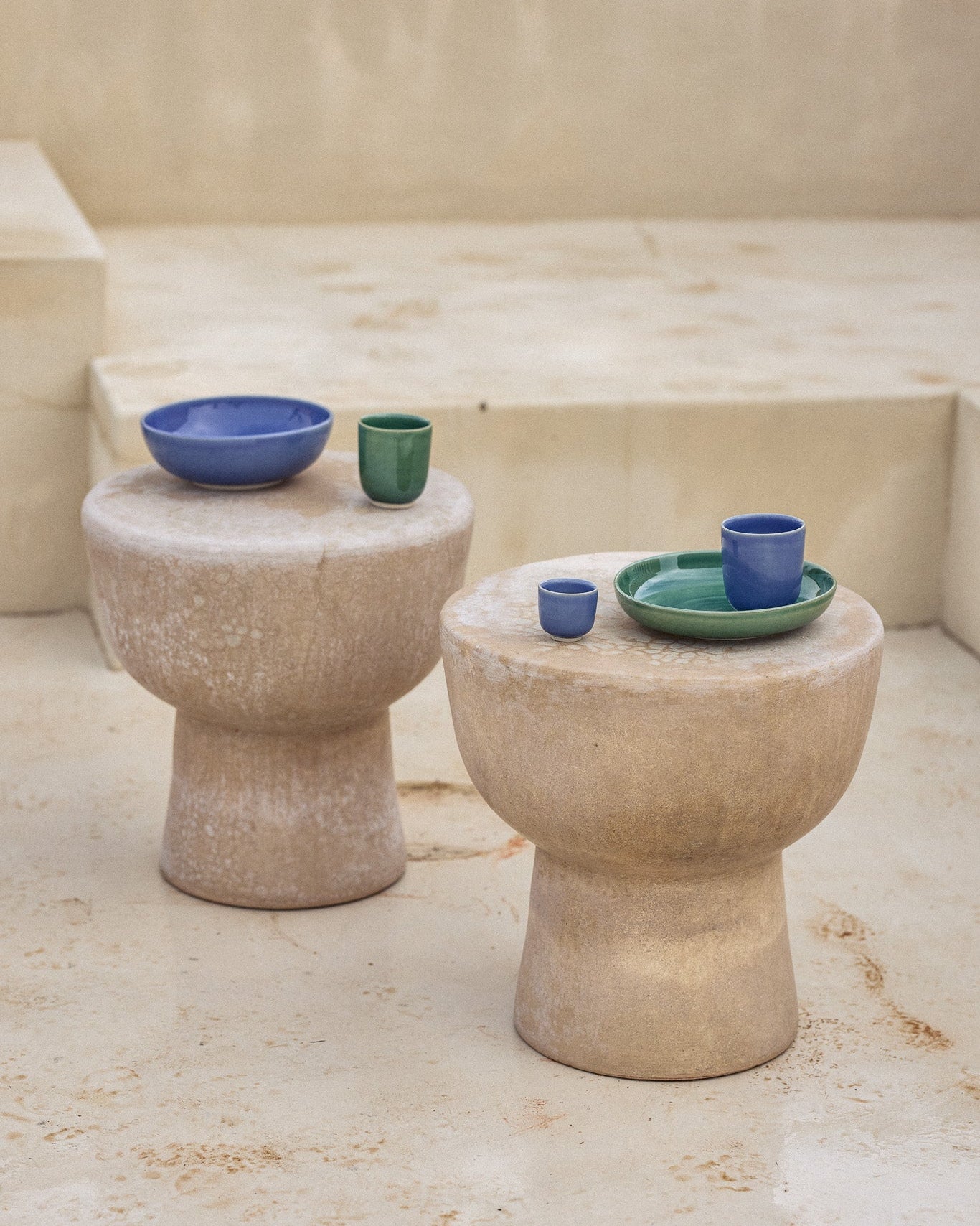 Zwei pilzförmige Beistelltische aus hellem Naturstein in einem mediterranen Außenbereich. Auf dem linken Tisch stehen eine blaue Keramikschüssel und ein grüner Keramikbecher, auf dem rechten ein grüner Teller mit einem blauen Becher und einem kleinen grünen Schälchen. Die leuchtenden Farben der glasierten Keramik setzen einen lebhaften Kontrast zur rauen, sandsteinfarbenen Oberfläche der Tische