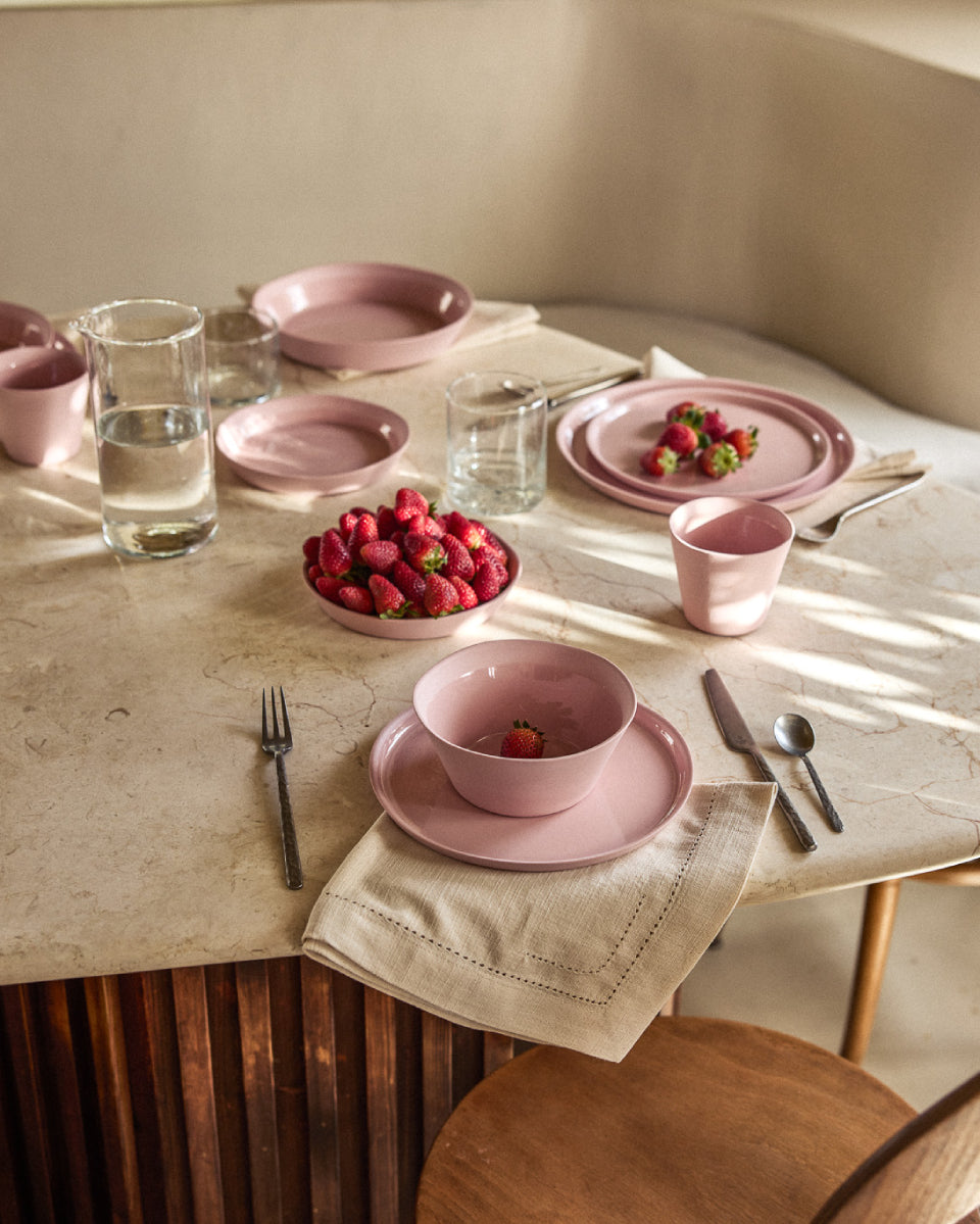 Sommerlich gedeckter runder Marmortisch mit rosafarbenem Keramikgeschirr. Eine große Schüssel ist gefüllt mit frischen Erdbeeren, weitere Erdbeeren liegen auf einem Teller. Im Vordergrund ein gedeckter Platz mit Schüssel auf Teller, Leinenserviette und dunklem Besteck. Klare Wassergläser und ein kleiner Becher ergänzen das harmonische Arrangement in Rosa- und Beigetönen. Der Tisch hat einen Holzunterbau, im Hintergrund ist ein Holzstuhl zu sehen.