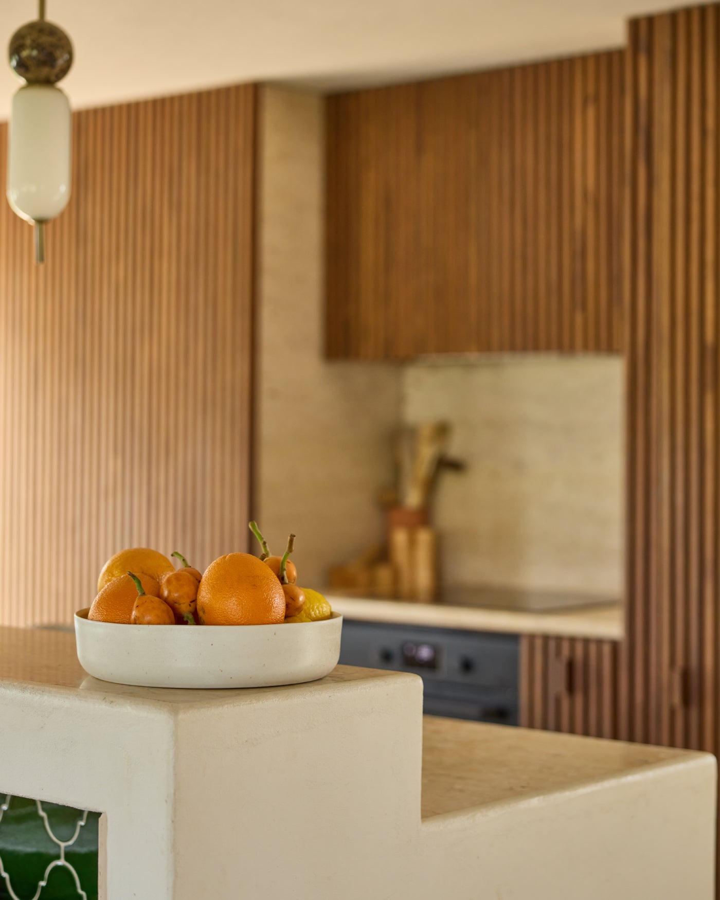 Cremefarbene Keramikschale mit Orangen und Kumquats auf einer hellen Steinarbeitsplatte. Im Hintergrund eine moderne Küche mit gerillten Holzfronten, blaugrauen Unterschränken und einer Messingarmatur. Links eine dekorative Pendelleuchte mit Glasschirm.