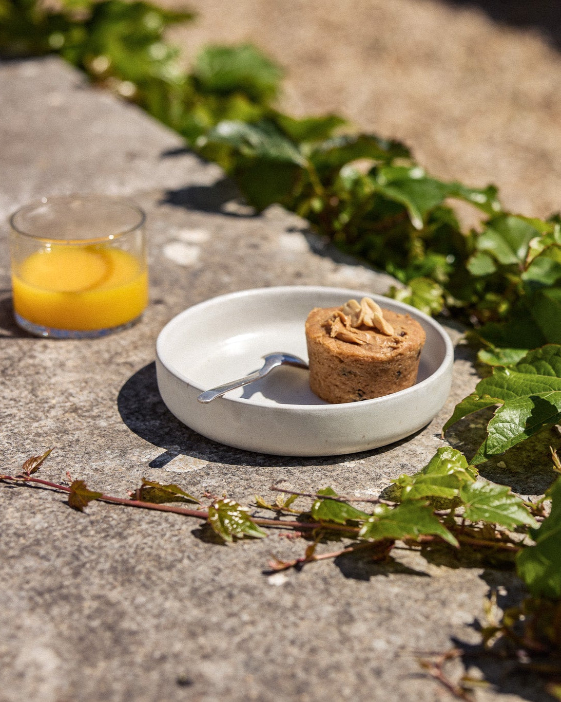 Cremefarbene Keramikschale mit einem kleinen Nuss-Muffin mit Erdnussbutter-Topping und einem Silberlöffel, auf einer Natursteinoberfläche im Sonnenlicht. Daneben ein Glas Orangensaft, umgeben von rankenden Efeuzweigen.
