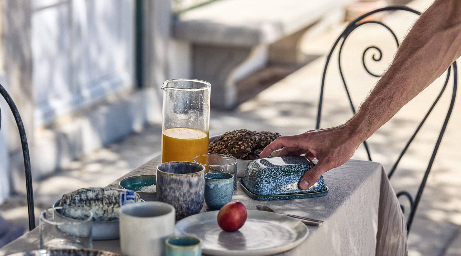 Eine Hand greift nach einer blau-glasierten Keramik-Butterdose auf einem sommerlich gedeckten Frühstückstisch im Freien. Auf dem leinenbedeckten Tisch stehen eine Glaskaraffe mit frisch gepresstem Orangensaft, mehrere Keramikbecher und -schüsseln in Blau- und Grautönen mit Reaktionsglasur, ein Teller mit dunklem Körnerbrot, eine rote Nektarine sowie weiteres Geschirr. Im Hintergrund sind schmiedeeiserne Stühle und eine helle mediterranee Architektur zu sehen.