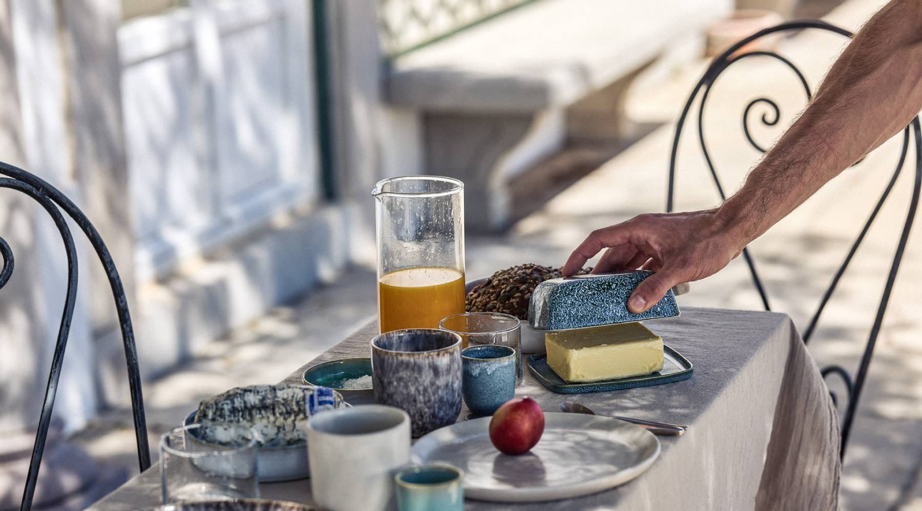 Hand greift nach einer blauen Keramik-Butterdose auf einem sonnigen Außentisch mit grauem Leinentischtuch. Auf dem Tisch stehen verschiedene blaue und graue Keramikbecher und -schalen, ein Glaskrug mit Orangensaft, eine Nektarine auf einem Teller sowie ein Käsestück. Beidseitig schwarze schmiedeeiserne Stühle, im Hintergrund eine helle Hausfassade.