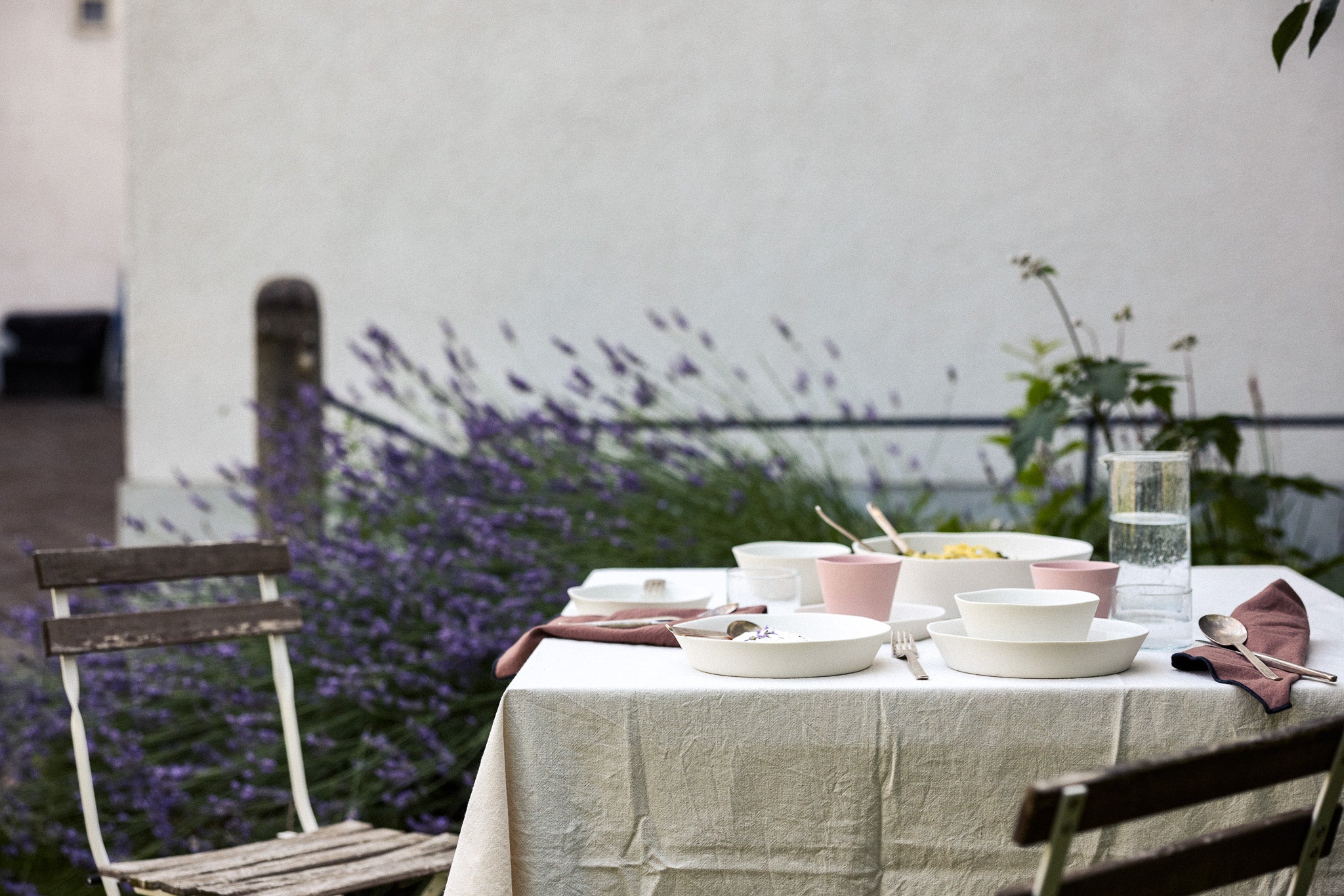 Gedeckter Gartentisch mit naturbeigem Leinentischtuch, weißen Keramikschalen und rosafarbenen Bechern, mauvefarbenen Stoffservietten, silbernem Besteck und einem Wasserkrug. Im Hintergrund blühender Lavendel vor einer weißen Hauswand sowie verwitterte Metallklappstühle.