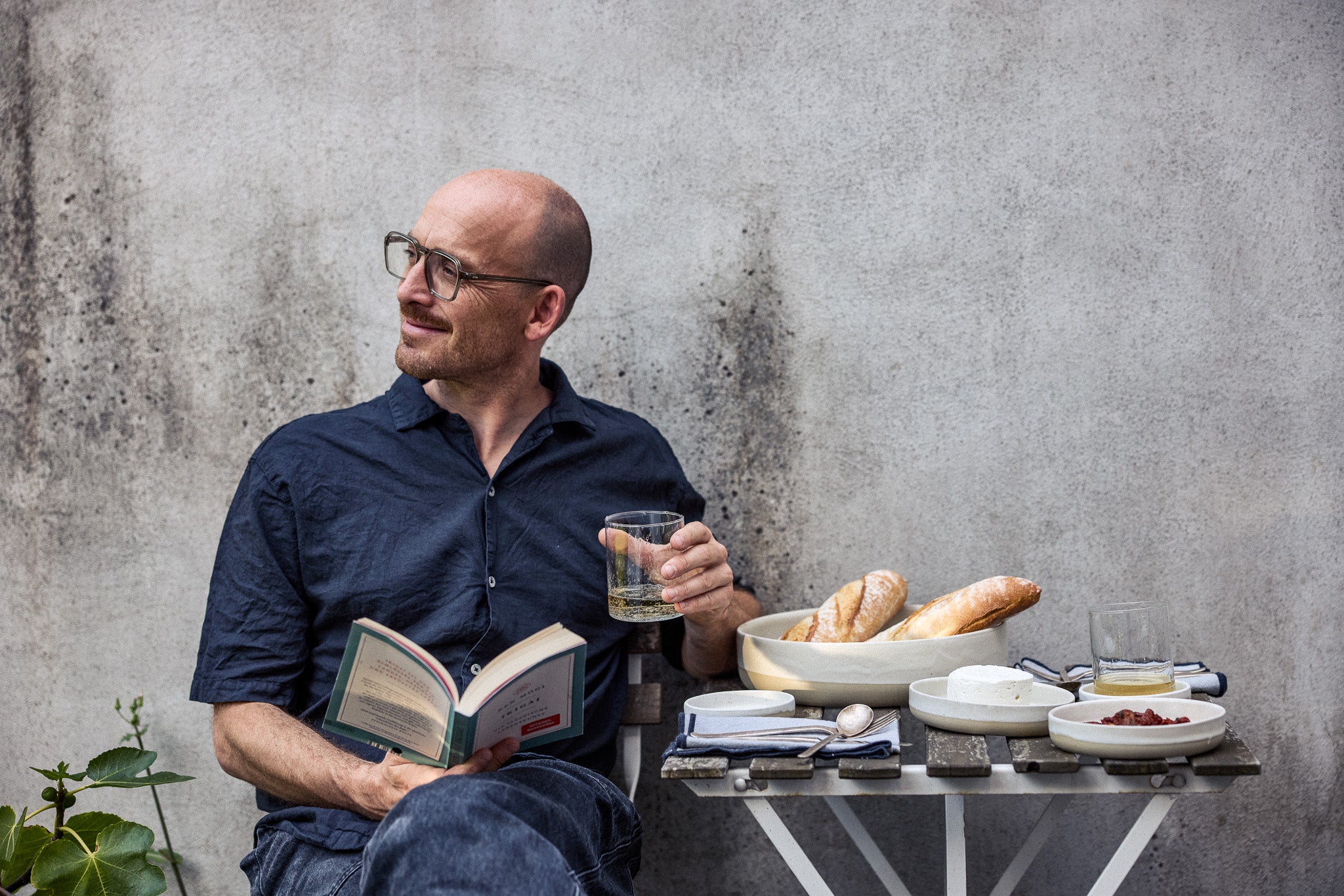 Mann mit Brille sitzt entspannt mit einem Buch und einem Glas Weißwein vor einer verwitterten Betonwand. Neben ihm ein Klapptisch mit cremefarbenem Keramikgeschirr, Baguettes, Frischkäse, getrockneten Tomaten und Besteck auf blau-weißen Stoffservietten.