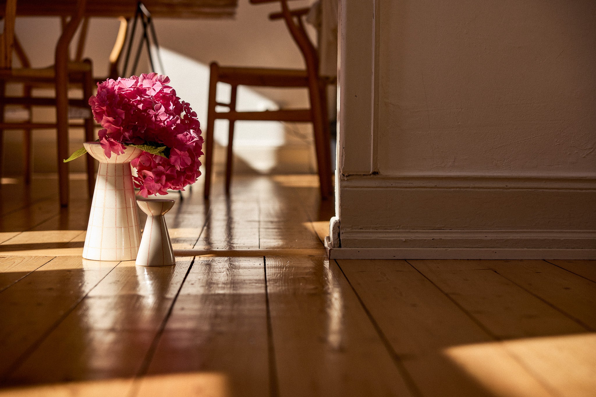 Eine schlanke, taillierte Keramikvase in Weiß mit einem üppigen Strauß leuchtend pinkfarbener Hortensien steht auf einem warmen Holzdielenboden im goldenen Gegenlicht. Die Vase befindet sich neben einem Türrahmen, im unscharfen Hintergrund sind Holzstühle eines Speisezimmers zu erkennen