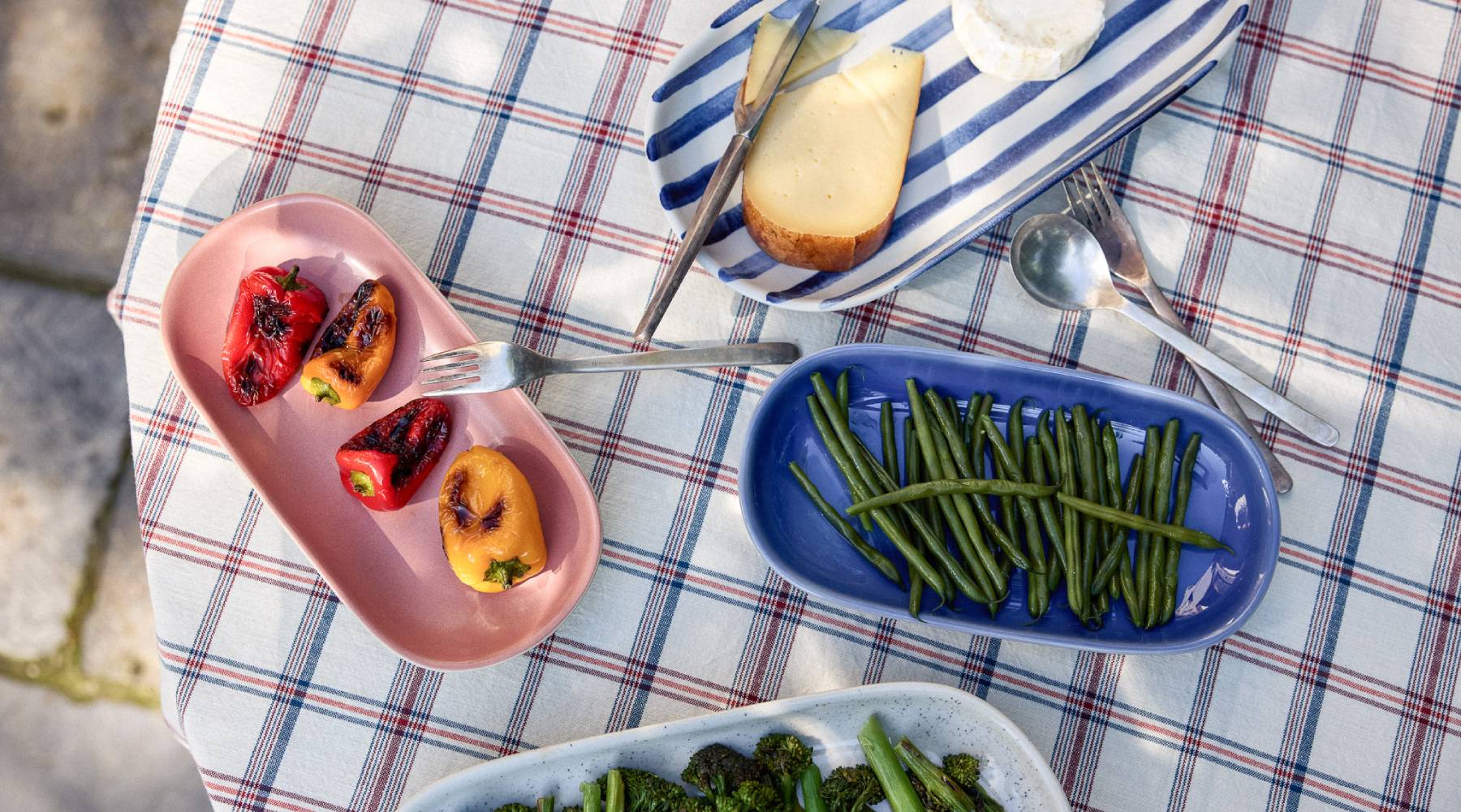 Vogelperspektive auf einen sommerlich gedeckten Tisch mit einem rot-weiß-blau karierten Tischtuch. Auf ovalen Serviertellern werden verschiedene Speisen präsentiert: gegrillte bunte Paprika auf einem rosafarbenen Teller, grüne Bohnen auf einem blauen Teller, gedünsteter Brokkoli auf einem weiß gesprenkelten Teller sowie überbackenes Brot auf einem blau-weiß gestreiften Teller