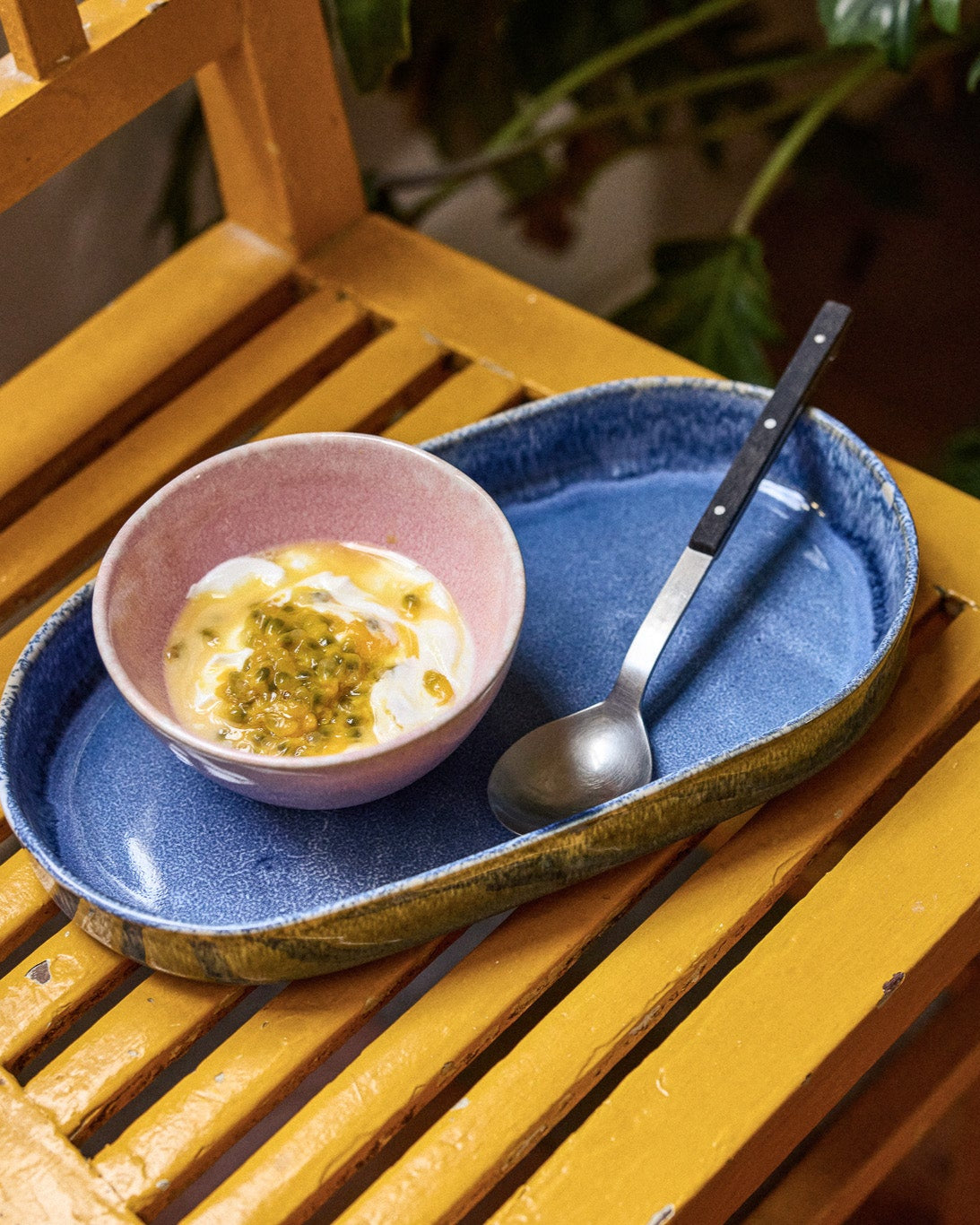 Rosa Keramikschale mit Joghurt und frischer Passionsfrucht auf einem ovalen blauen Keramiktablett mit gelbem Rand, daneben ein silberner Löffel. Das Arrangement liegt auf einem leuchtend gelben Holzstuhl, im Hintergrund grüne Pflanzen.