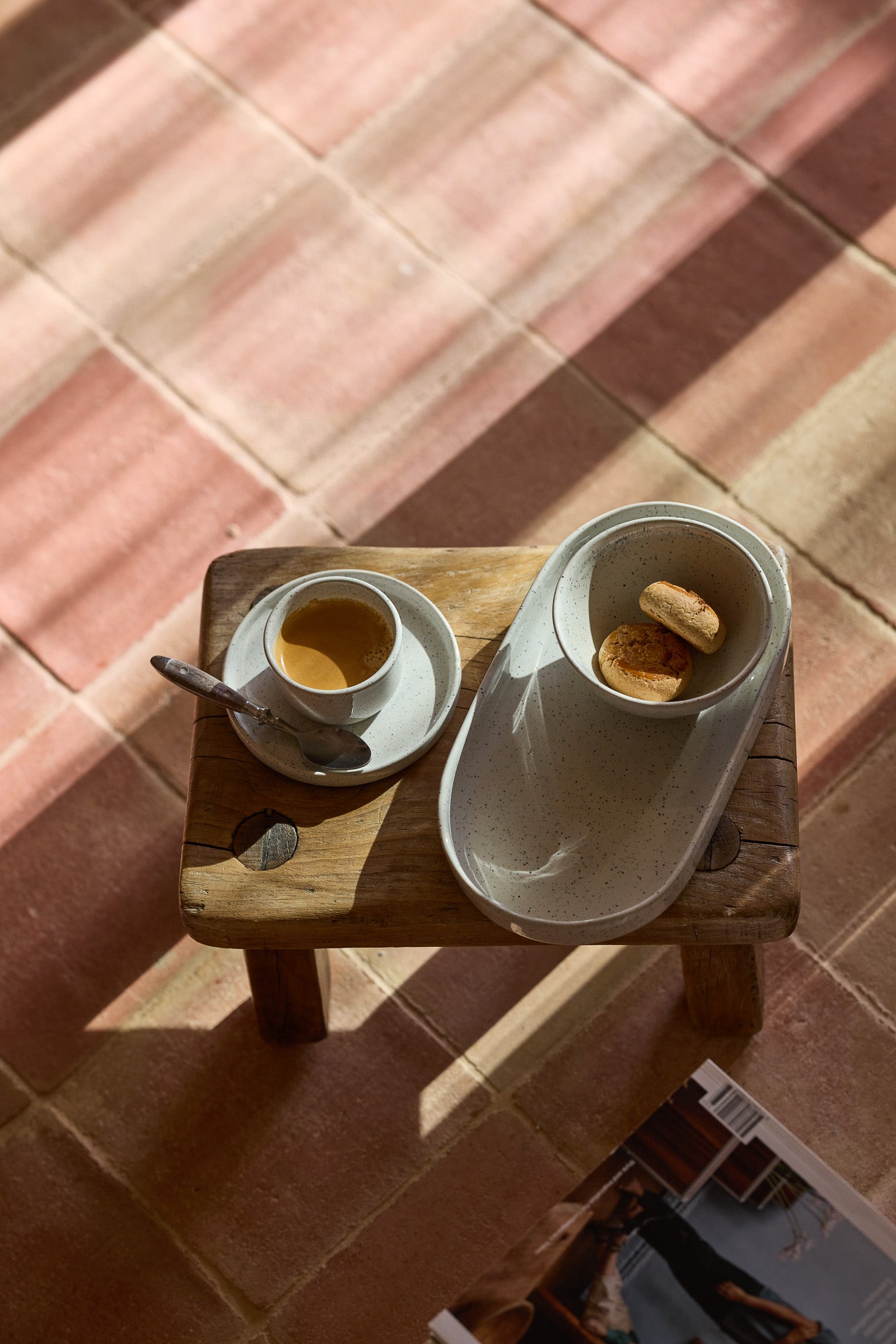 Kleiner rustikaler Holzhocker von oben mit einer gesprenkelten cremefarbenen Keramikespressotasse auf Unterteller mit Silberlöffel sowie einem ovalen Keramiktablett mit zwei kleinen Brioche-Brötchen. Auf rosafarbenem Terrakottafliesenboden im Sonnenlicht.
