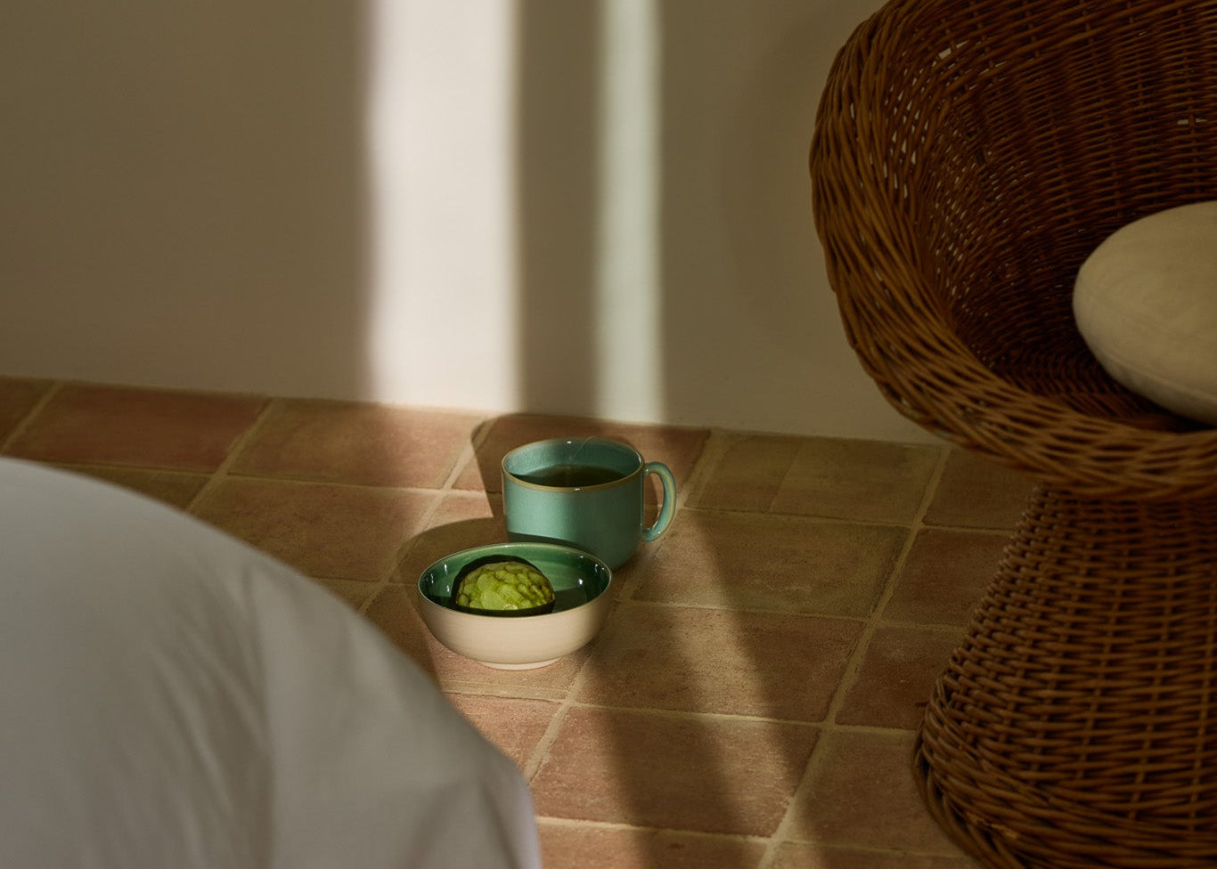 Türkisfarbene Keramiktasse mit Tee und eine cremefarbene Schale mit einer halben Avocado direkt auf einem Terrakottafliesenboden im warmen Sonnenlicht. Rechts ein Rattansessel, links ein weißes Sofa angeschnitten.