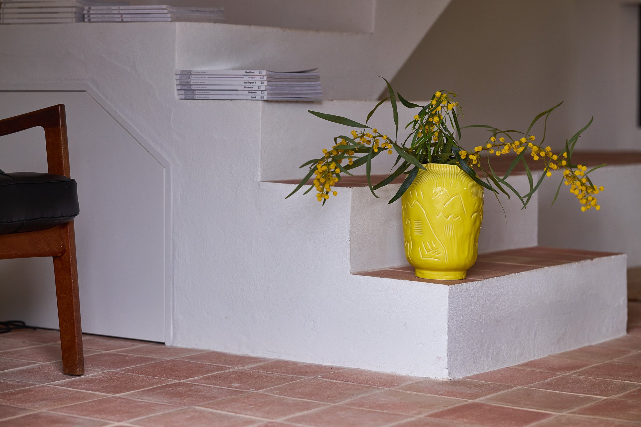 Eine leuchtend gelbe Keramikvase mit eingeritztem Reliefmuster steht auf einer weißen Treppenstufe mit Terrakottafliesen. In der Vase ein üppiger Strauß gelber Mimosen mit langen grünen Zweigen. Links ein Holzstuhl mit schwarzem Ledersitz, oben auf der Treppe ein Stapel Kunstkataloge oder Magazine. Die Szene strahlt eine lebendige, mediterrane Wohnatmosphäre aus.