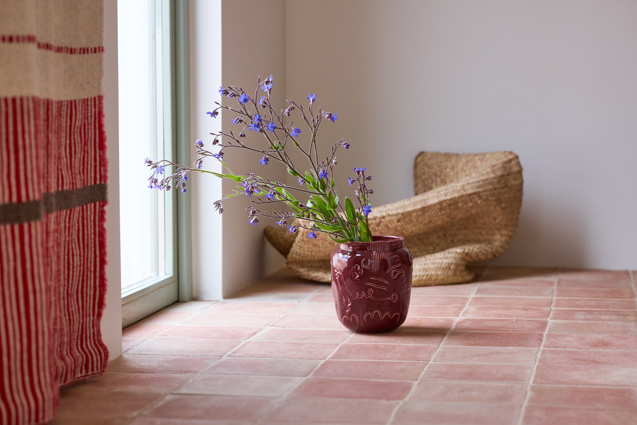 Eine bauchige, dunkelrote Keramikvase mit eingeritzten Linien und floralem Reliefmuster steht auf einem Terrakottafliesenboden vor einem hellen Fenster. In der Vase ein lockerer Strauß zarter blauer Wildblumen. Links ein rot-beige gestreifter Vorhang, rechts ein organisch geformter Korbsessel aus Naturfaser. Die Szene strahlt eine warme, mediterrane Wohnatmosphäre aus.