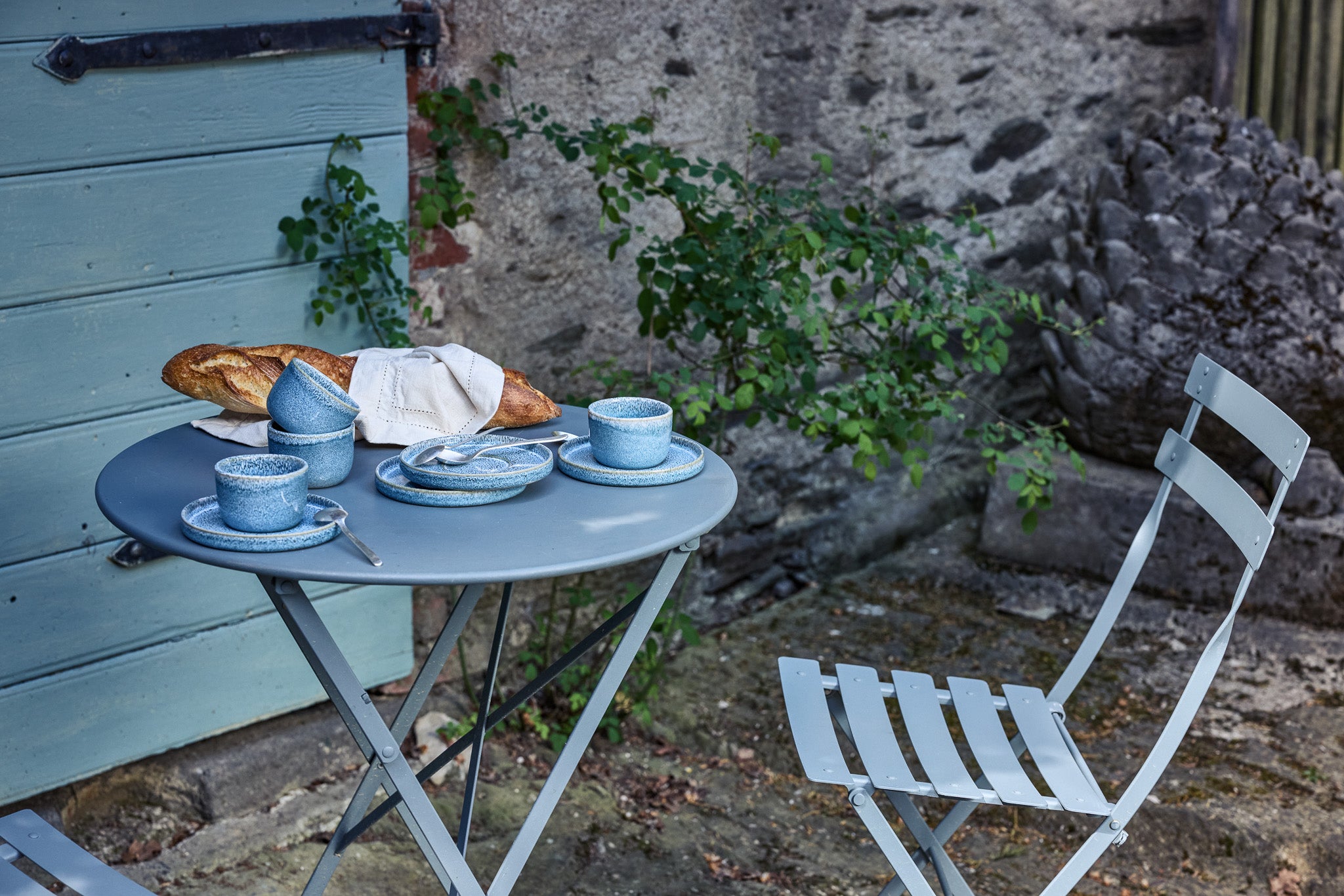 Grauer Klapptisch mit blauem Keramikgeschirr (Tassen, Schalen und Teller) und einem Baguette, aufgestellt auf einem Kopfsteinpflaster-Hof neben einer mintgrünen Holzwand und einer Natursteinmauer. Ein passender Klappstuhl steht daneben.