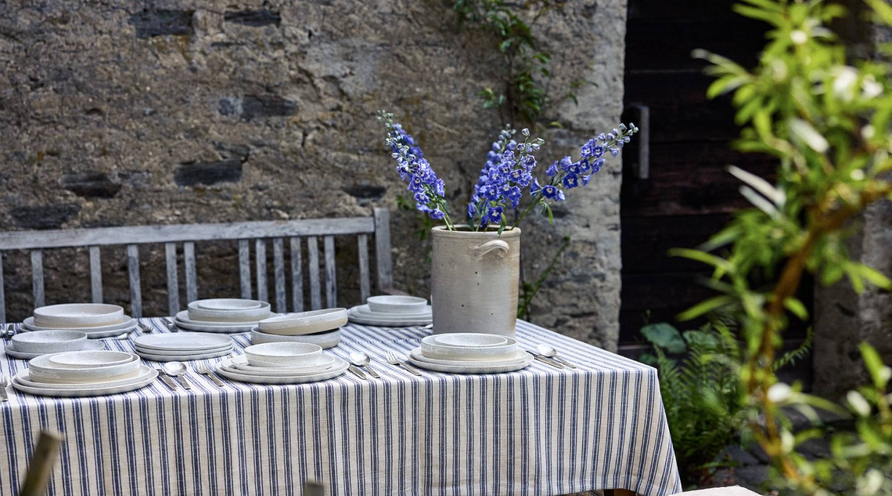 Gedeckter Outdoortisch mit blau-weiß gestreiftem Leinentischtuch, darauf cremefarbenes gesprenkeltes Keramikgeschirr (Teller und Schalen) mit silbernem Besteck. Als Tischdeko ein rustikaler Steingut-Krug mit blauen Rittersporn-Blüten. Im Hintergrund eine Natursteinmauer und eine verwitterte Holzbank, rechts üppiges Grün.