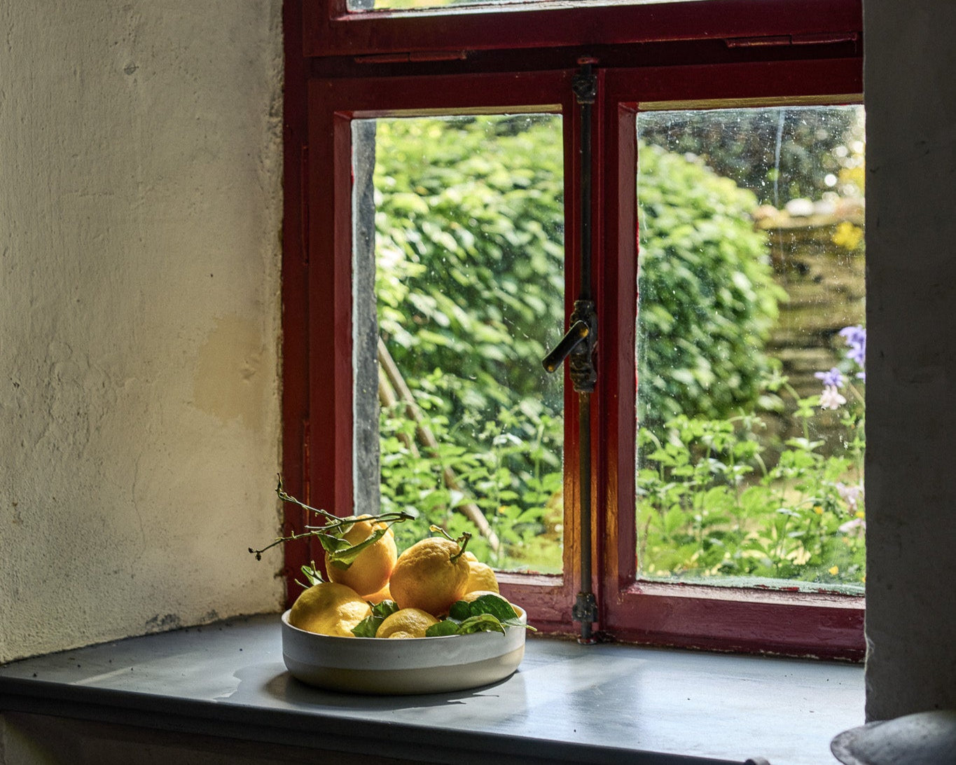 Eine flache Keramikschale mit frischen Zitronen und grünen Blättern auf einem Fensterbrett aus Stein. Das alte, rotgerahmte Fenster mit Sprossen ist leicht geöffnet und gibt den Blick auf einen üppig begrünten Garten frei. Die verwitterten Wände und das natürliche Licht verleihen der Szene einen malerisch-rustikalen Charakter