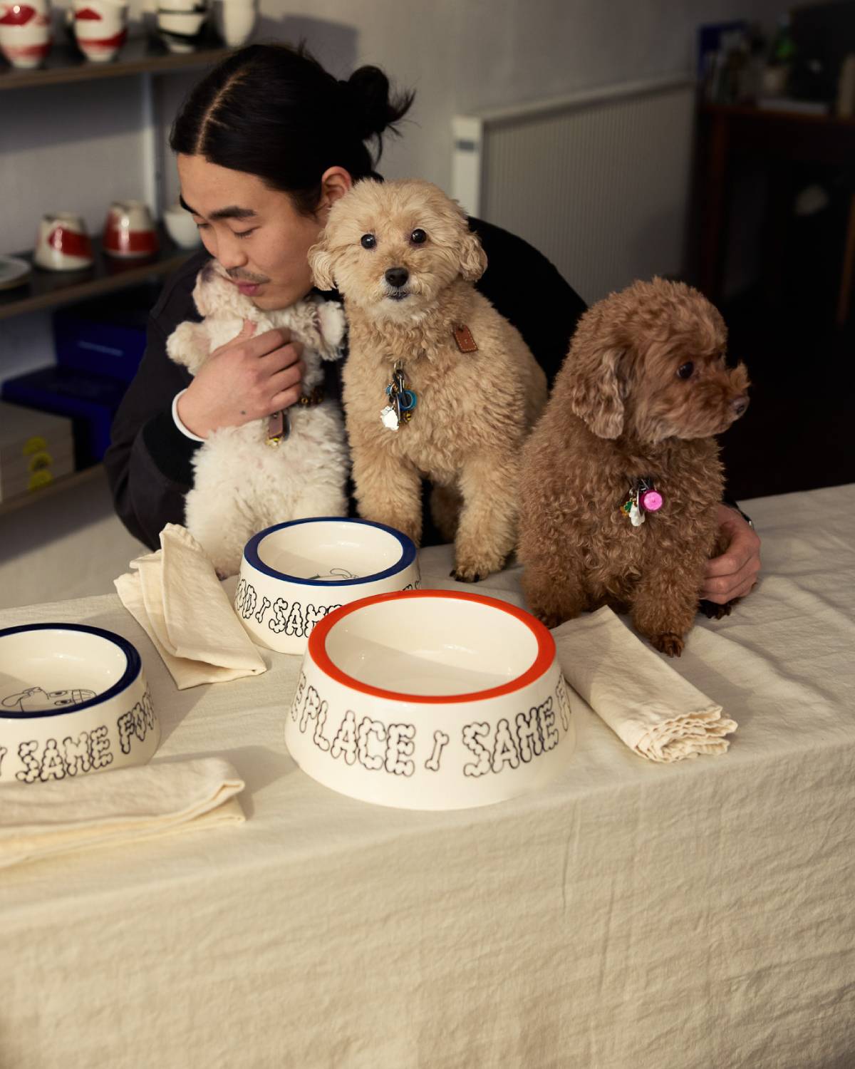Person mit zwei Hunden hinter einem Tisch mit weißen Keramiknäpfen mit blauem und rotem Rand und Aufschrift