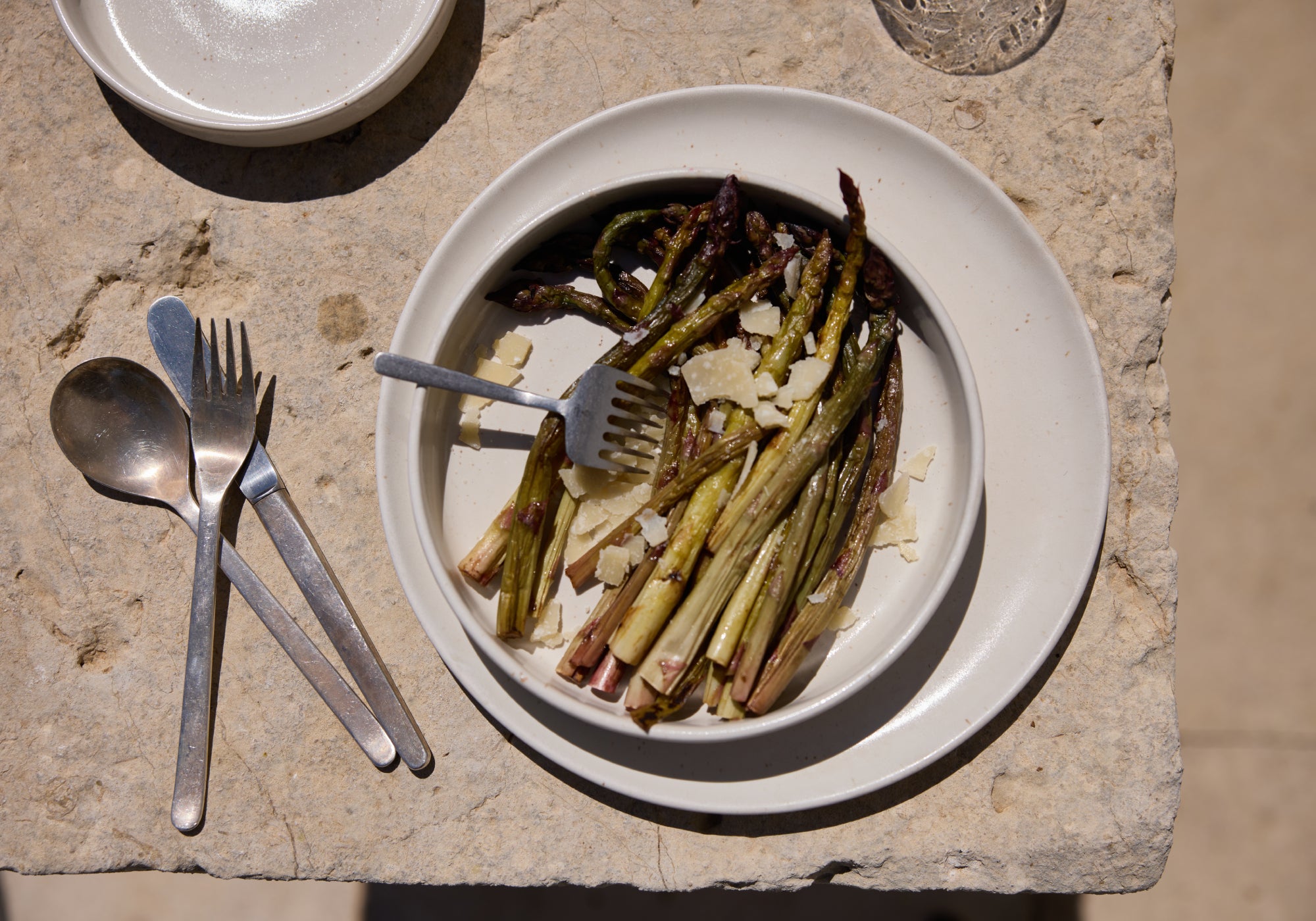 Gegrillter grüner Spargel mit Parmesanspänen auf einem cremefarbenen Keramikteller, serviert auf einer Natursteinplatte im Sonnenlicht. Daneben Gabel und Löffel aus Silber.