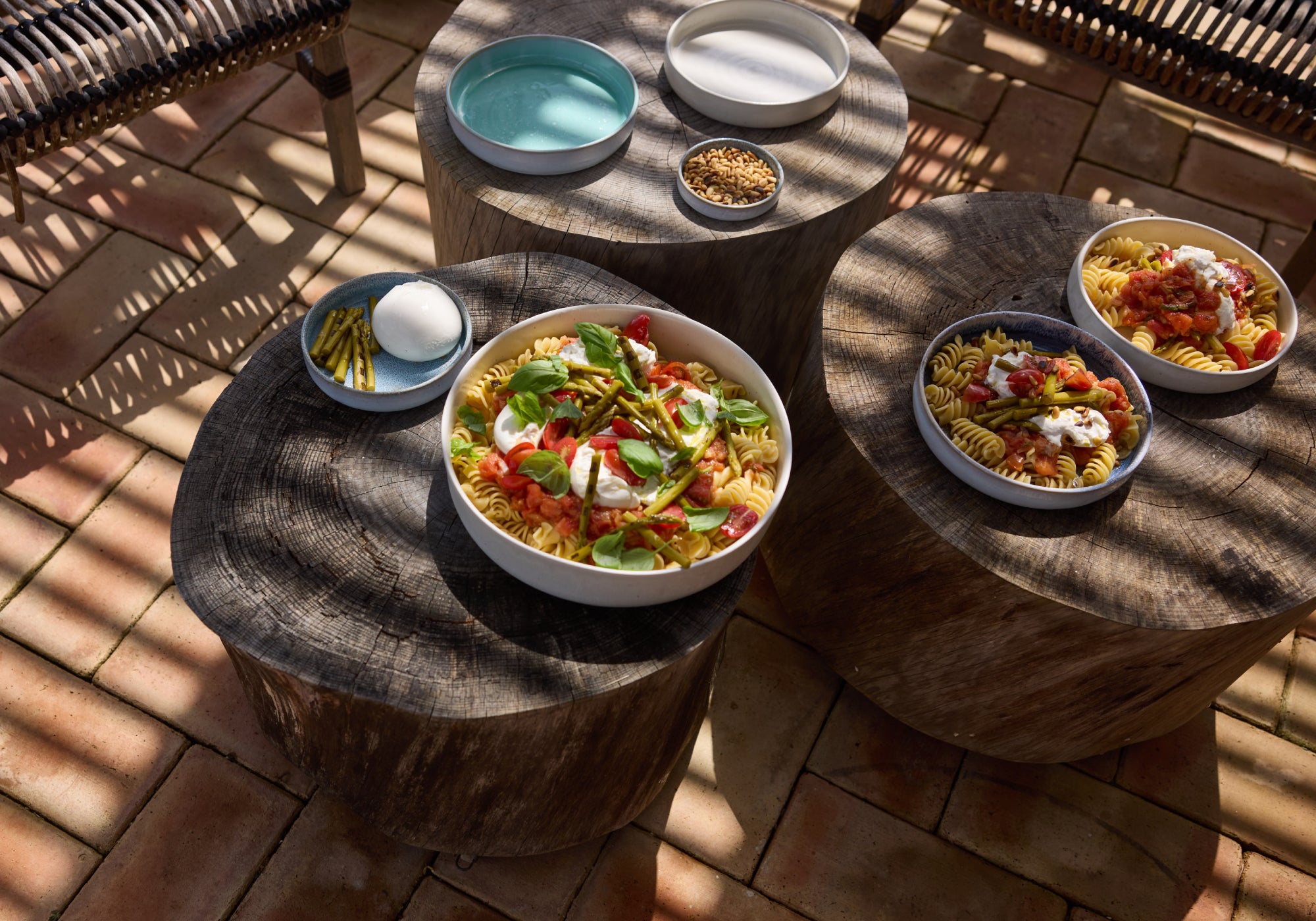 Rustikale Baumstumpf-Beistelltische auf einem Terrakotta-Fliesenboden, darauf weiße und blaue Keramikschalen mit Fusilli-Nudelsalat (Tomaten, grüne Bohnen, Burrata und Basilikum). Daneben ein blauer Teller mit Mozzarella und Bohnen sowie eine kleine Schale mit Pinienkernen und eine türkise Schale. Im Hintergrund Rattanmöbel im Schatten.