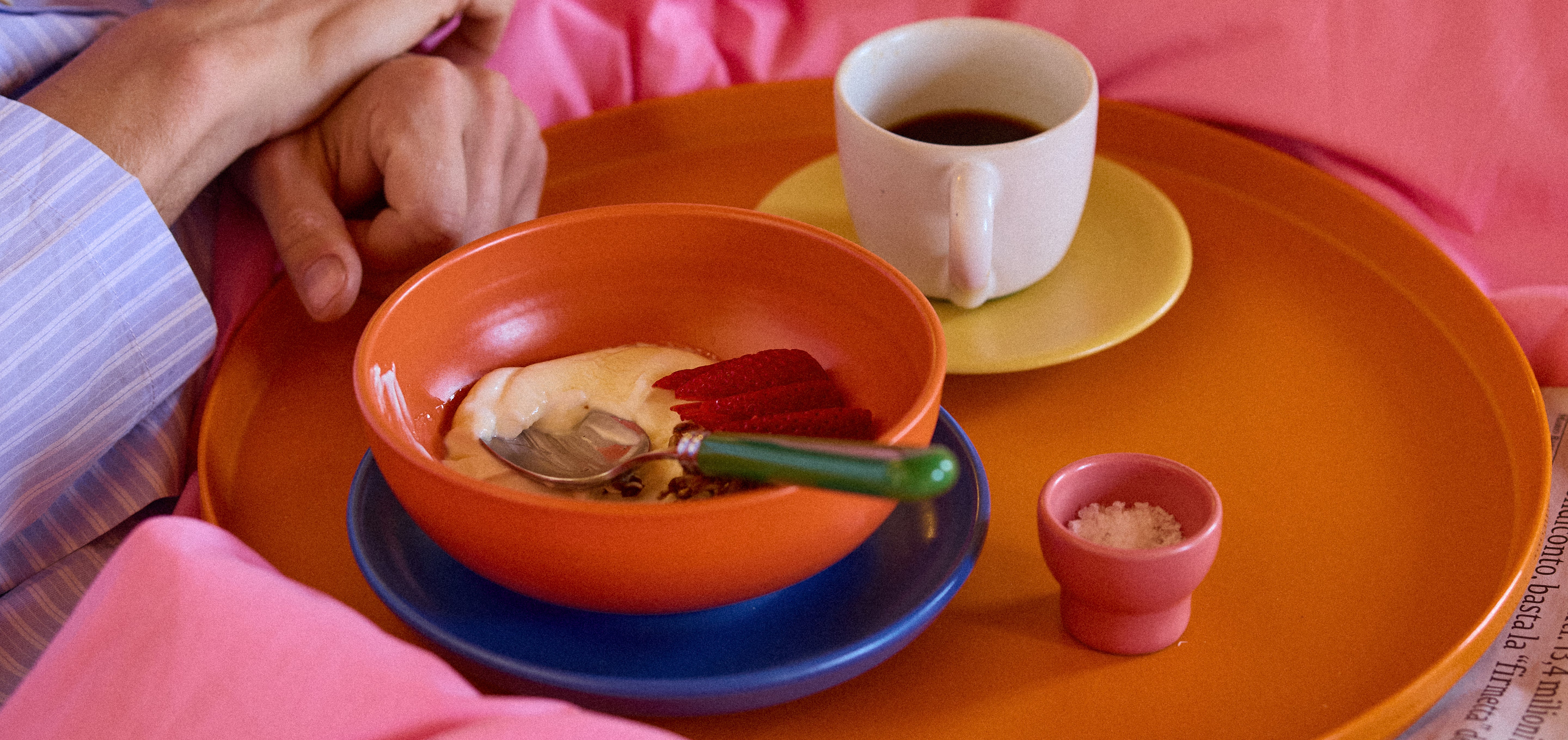 Frühstück im Bett auf einem runden orangefarbenen Tablett, umgeben von pinkfarbener Bettwäsche. Auf dem Tablett steht eine orangefarbene Keramikschüssel mit Joghurt und einem grün-stieligen Löffel auf blauem Unterteller, eine cremefarbene Kaffeetasse auf gelbem Teller sowie ein kleines rosafarbenes Keramikschälchen mit Zucker oder Salz. Eine Hand im gestreiften Pyjama greift ans Tablett.