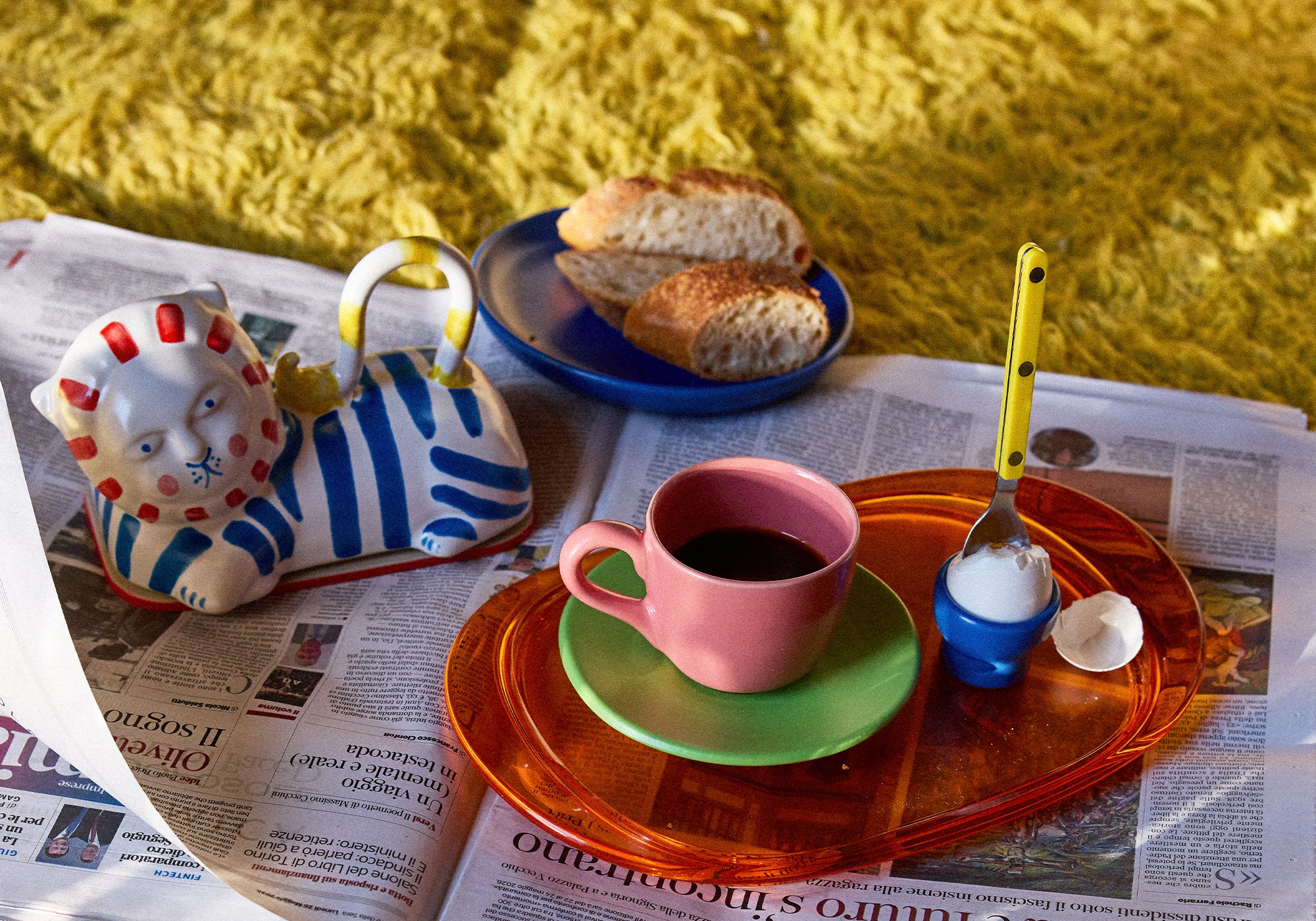 Verspieltes Frühstücksstillleben auf aufgefächerten italienischen Zeitungen vor einem gelben Flauschteppich. Auf einem orangefarbenen Glastablett stehen eine rosafarbene Kaffeetasse auf grüner Untertasse, ein blauer Eierbecher mit gekochtem Ei und ein gelb-gepunkteter Löffel. Daneben eine blau-weiß gestreifte Keramik-Teekanne in Löwenform mit roten Details. Im Hintergrund ein blauer Teller mit aufgeschnittenen Baguettescheiben.