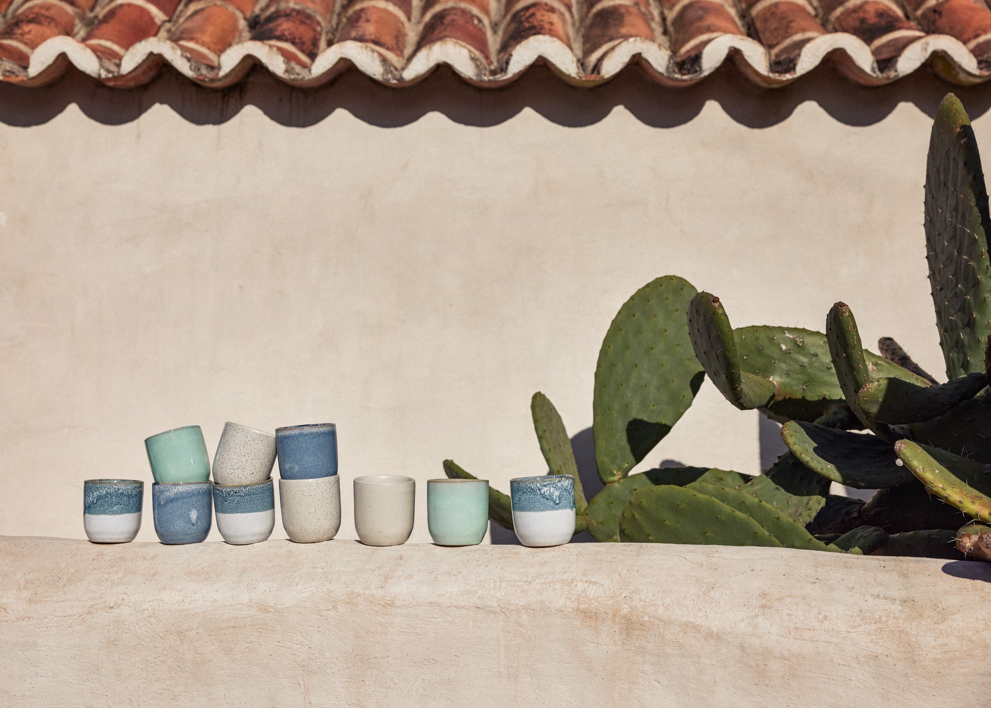 Eine Reihe handgefertigter Keramikbecher in verschiedenen Größen und Farben – Blau, Mint, Beige und Naturweiß mit zweifarbiger Glasur – auf einer hellen Steinmauer im Freien. Rechts im Bild ein großer Feigenkaktus. Im Hintergrund eine mediterrane Lehmwand mit traditionellem Ziegeldach, im hellen Sonnenlicht.