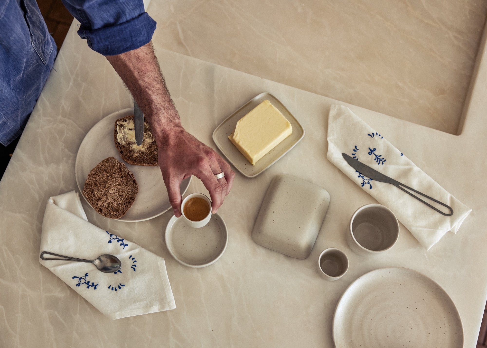 Vogelperspektive auf einen hellen Frühstückstisch. Eine Hand mit Ring greift nach einer kleinen Espressotasse auf Untertasse aus naturfarben gesprenkelter Keramik. Daneben ein Teller mit Körnerbrotscheiben, eine Butterdose mit frischer Butter, eine geschlossene quadratische Keramikdose sowie kleine Becher und ein Teller in gleichem Steinzeugton. Zwei weiße Leinenservietten mit blauer Stickerei und je einem Besteckteil vervollständigen das minimalistische Arrangement
