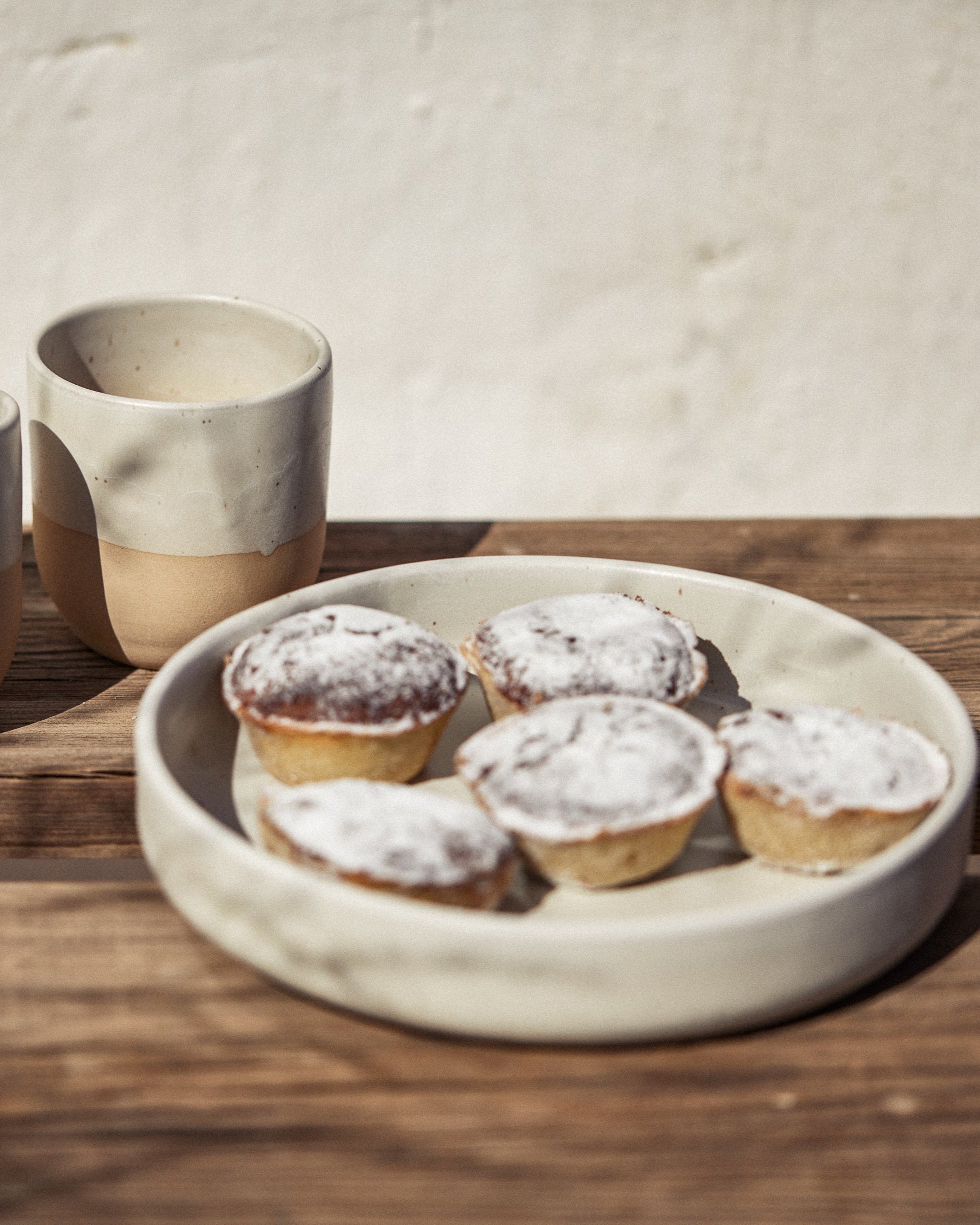 Cremefarbene Keramikschale mit vier mit Puderzucker bestäubten Mini-Törtchen auf einem Holztisch im Sonnenlicht, daneben ein zweitoniger Keramikbecher mit weißer Glasur und naturbelassenem Ton vor heller Wand.