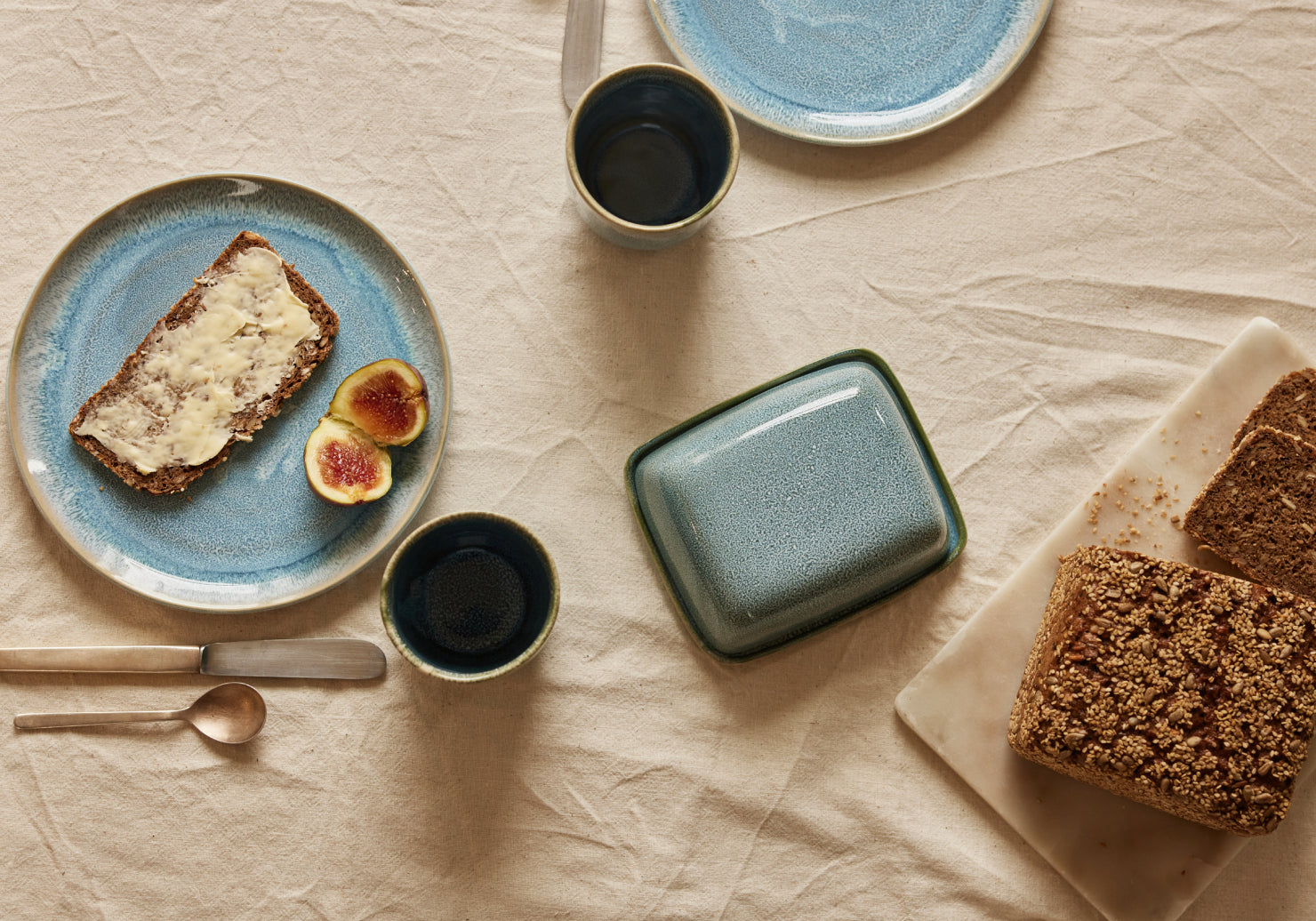 Vogelperspektive auf ein Frühstücksarrangement auf cremefarbenem Leinenstoff. Auf einem blau-glasierten Keramikteller liegt eine gebutterte Scheibe Körnerbrot mit zwei halbierten Feigen. Daneben eine türkis-grüne Keramik-Butterdose, zwei kleine dunkle Keramikbecher, ein weiterer blauer Teller im Hintergrund sowie aufgeschnittenes Körnerbrot auf einem weißen Marmorschneidebrett. Goldenes Besteck liegt links im Bild.