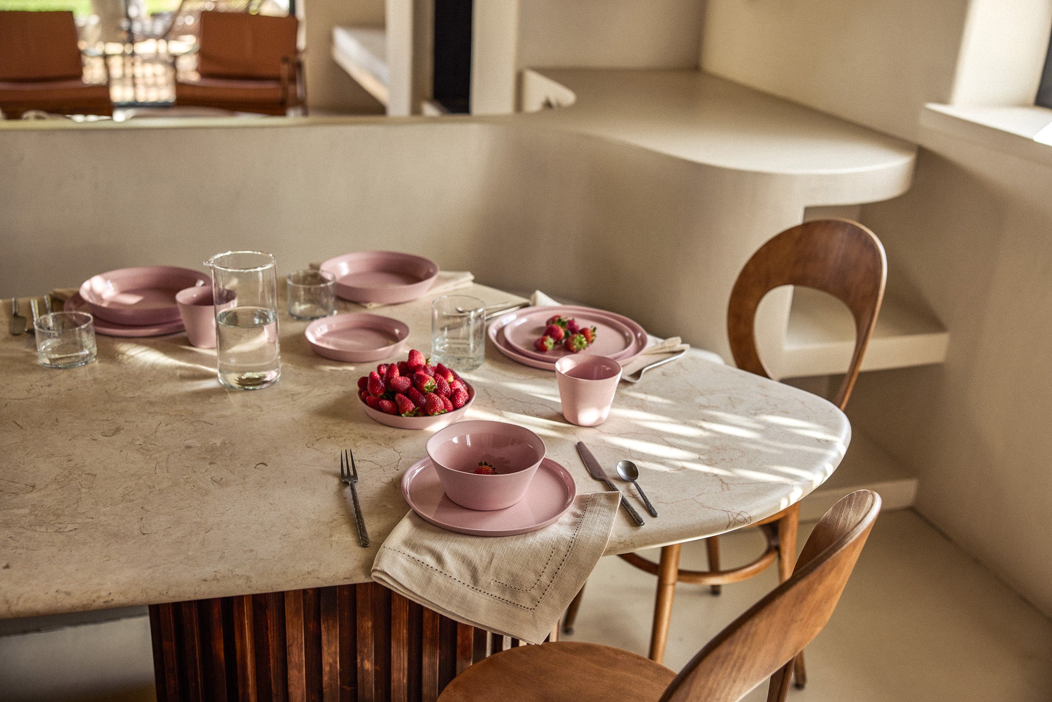 Ovaler Marmortisch mit Holzunterbau in einem warmen, minimalistischen Interieur, gedeckt mit rosafarbenem Keramikgeschirr. Eine große Schüssel ist gefüllt mit frischen Erdbeeren, weitere Erdbeeren liegen auf einem Teller. Klare Wassergläser, ein rosafarbener Becher und Leinenservietten mit Hohlsaumstickerei ergänzen das Arrangement. Zwei Holzstühle mit geschwungener Rückenlehne stehen am Tisch, im Hintergrund eine organisch geformte Wandnische.