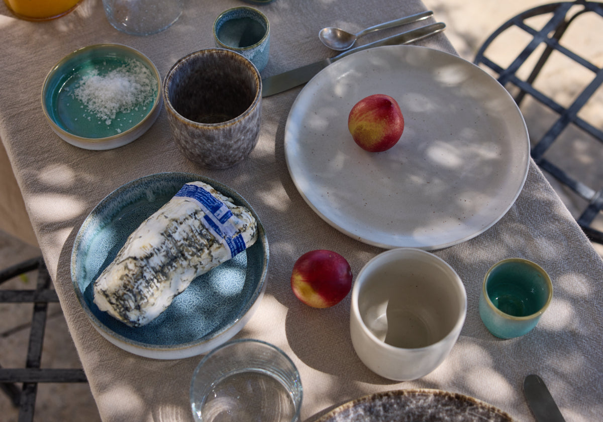 Outdoortisch von oben mit grauem Leinentischtuch, darauf verschiedene Keramikgefäße in Blau, Grün und Weiß. Auf einem blauen Teller ein Ziegenrolle-Käse, auf einem weißen Teller eine Nektarine, eine weitere Nektarine liegt lose auf dem Tisch. Eine türkise Schale mit Meersalz, Gläser und Besteck vervollständigen das sommerliche Frühstücksarrangement.