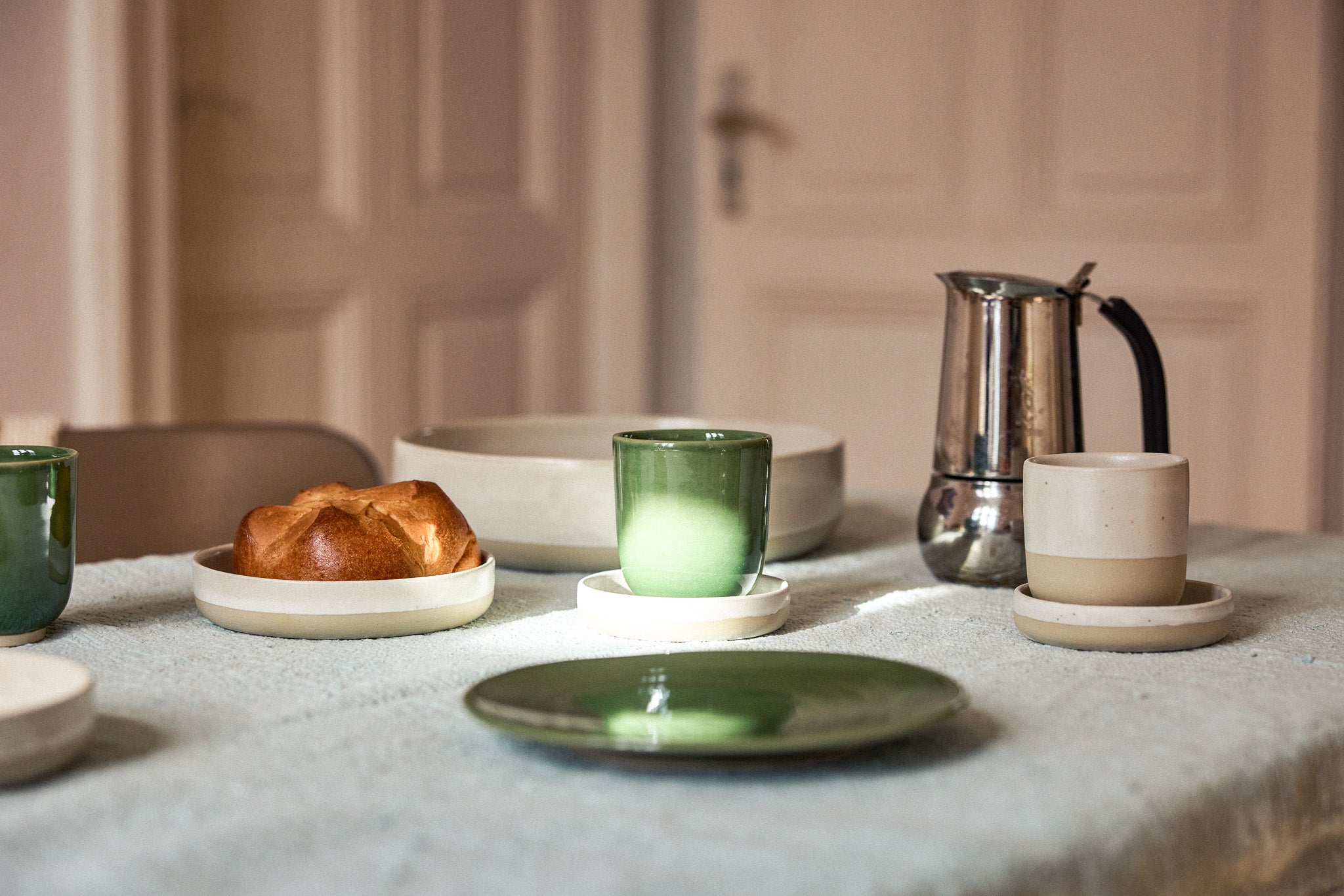 Frühstückstisch mit grauem Leinentischtuch, darauf grüne Keramikteller und -becher mit Verlaufsglasur auf cremefarbenen Untersetzern, ein cremefarbener Becher, eine große Schale, ein goldbraunes Brioche-Brötchen sowie eine glänzende Edelstahl-Mokakanne. Im Hintergrund eine rosafarbene Holztür.