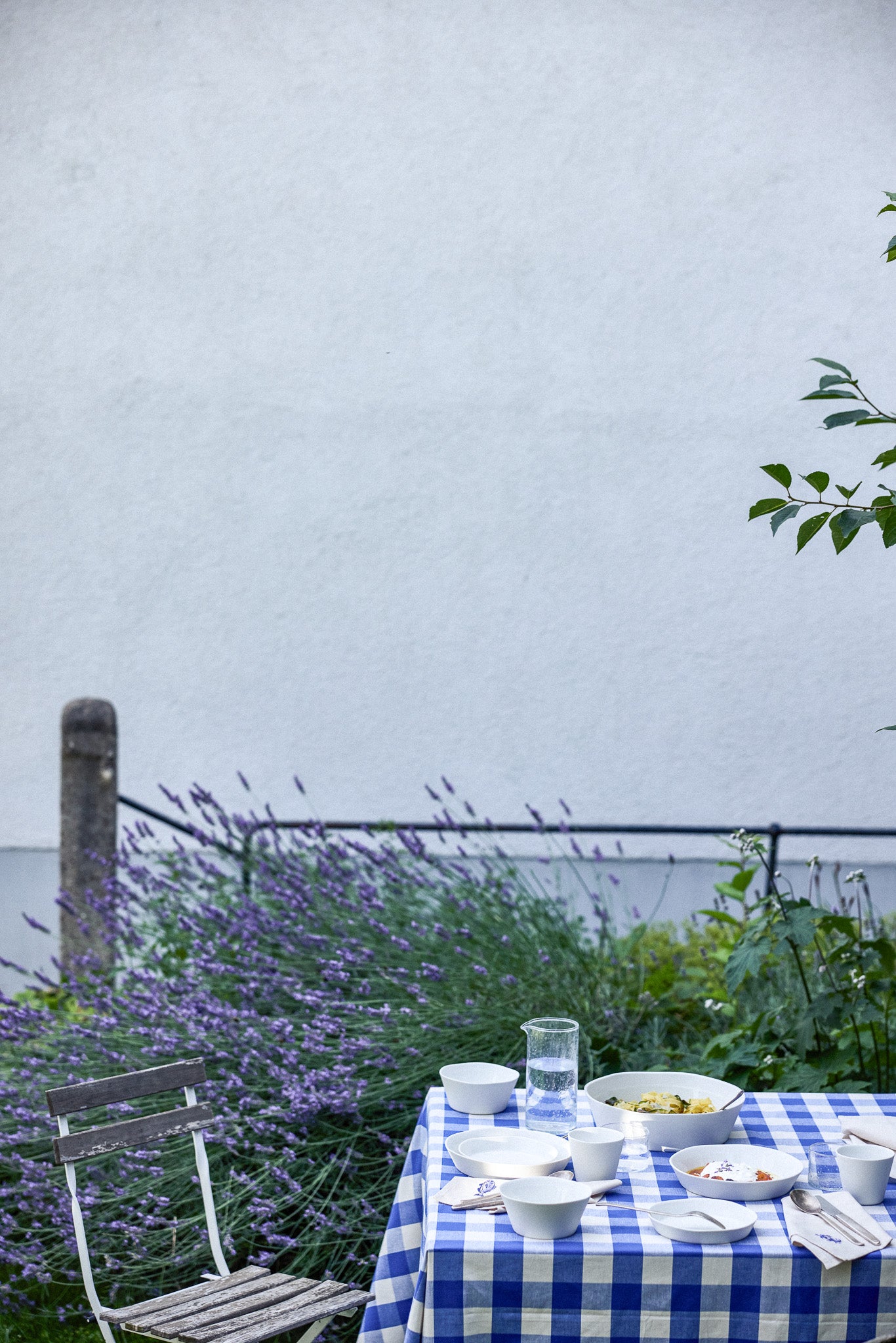 Gedeckter Gartentisch mit blau-weiß kariertem Tischtuch, darauf weiße Keramikschalen mit Speisen, ein Glaskrug mit Wasser und Besteck. Im Hintergrund blühender Lavendel vor einem schwarzen Metallzaun und einer weißen Hauswand, rechts ein Holzklappstuhl.