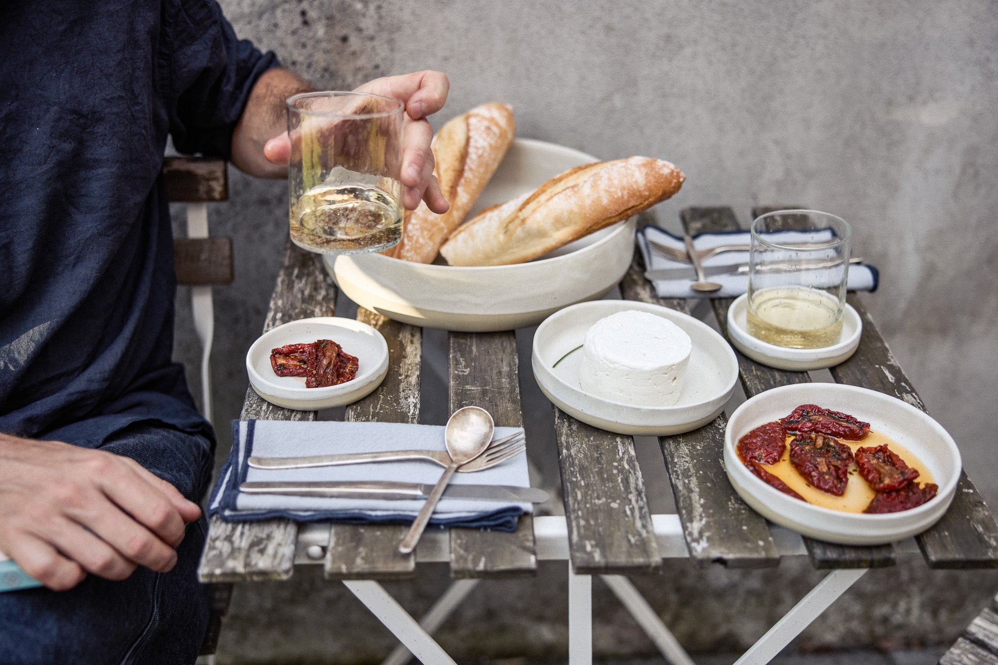 Person greift nach einem Glas Weißwein auf einem verwitterten Holzklapptisch mit cremefarbenem Keramikgeschirr: eine große Schale mit Baguettes, ein runder Frischkäse auf einem Teller, getrocknete Tomaten auf zwei kleinen Tellern sowie ein zweites Weinglas. Dazu blau-weiße Stoffservietten mit Besteck.
