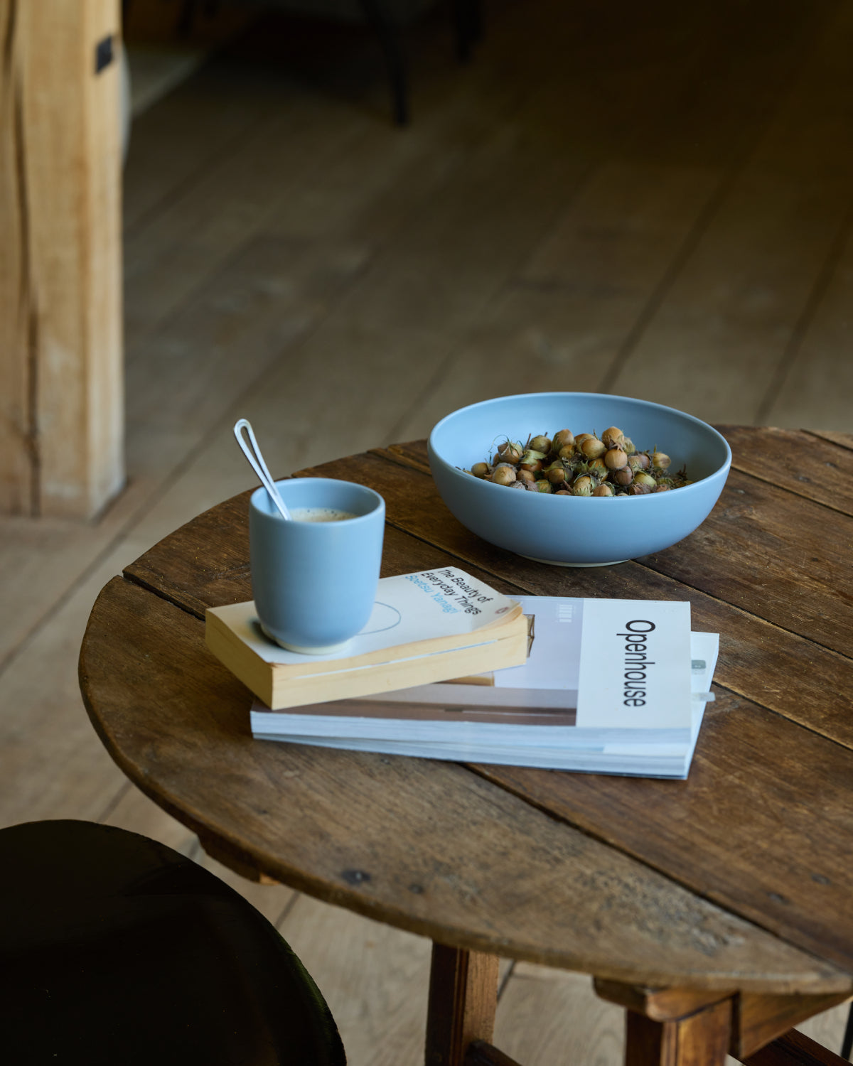Rustikaler runder Holztisch mit einer hellblauen Keramikschale voller Haselnüsse, einem blauen Keramikbecher mit Kaffee und einem weißen Löffel, gestapelt auf Büchern und Magazinen – darunter das „Openhouse"-Magazin. Im Hintergrund ein Holzdielenboden.