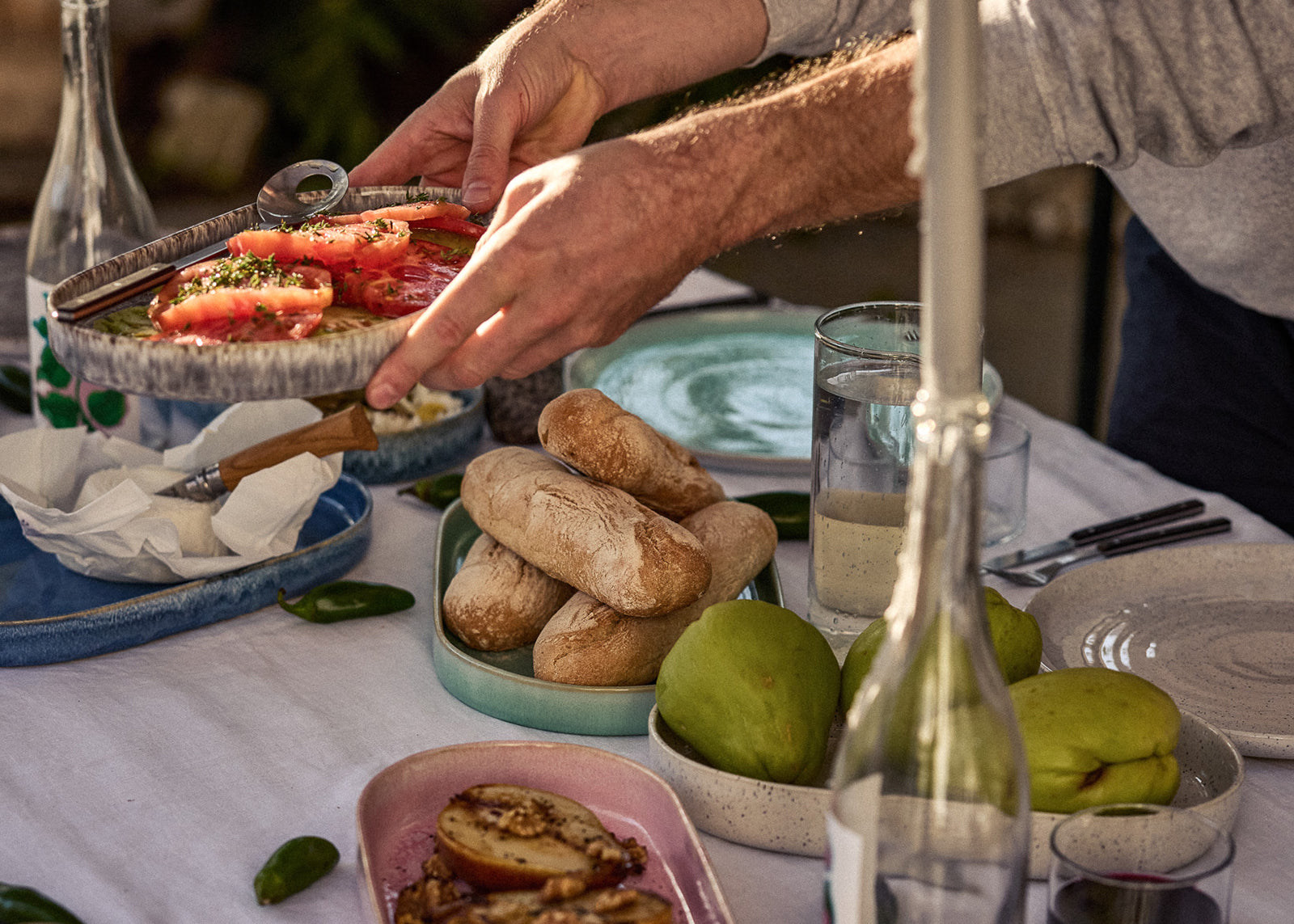 Eine Person reicht bei einem gedeckten Außentisch eine Keramikschale mit frischen Tomatenscheiben und Kräutern weiter. Auf dem Tisch stehen weitere Speisen: rustikale Brötchen auf einem türkis-grünen Teller, grüne Birnen in einer gesprenkelten Schüssel, karamellisierte Früchte auf einem rosafarbenen Teller sowie Käse auf einem blauen Teller. Glaskaraffen und Trinkgläser vervollständigen das sommerliche Tafelbild.