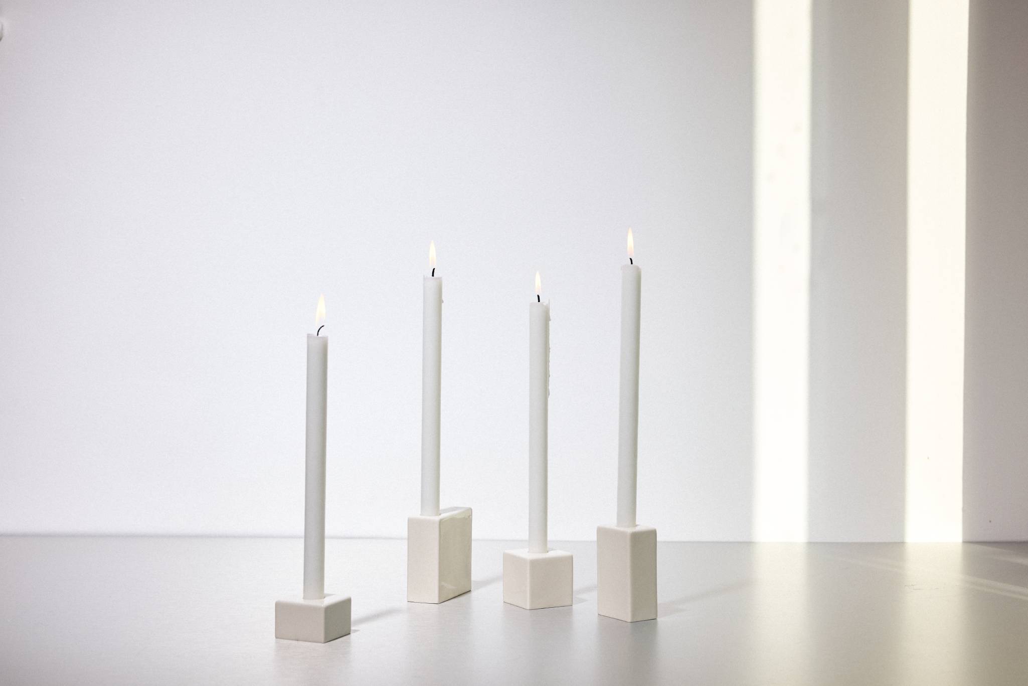 Drei brennende weiße Stabkerzen in minimalistischen quadratischen Keramikhaltern vor weißem Hintergrund