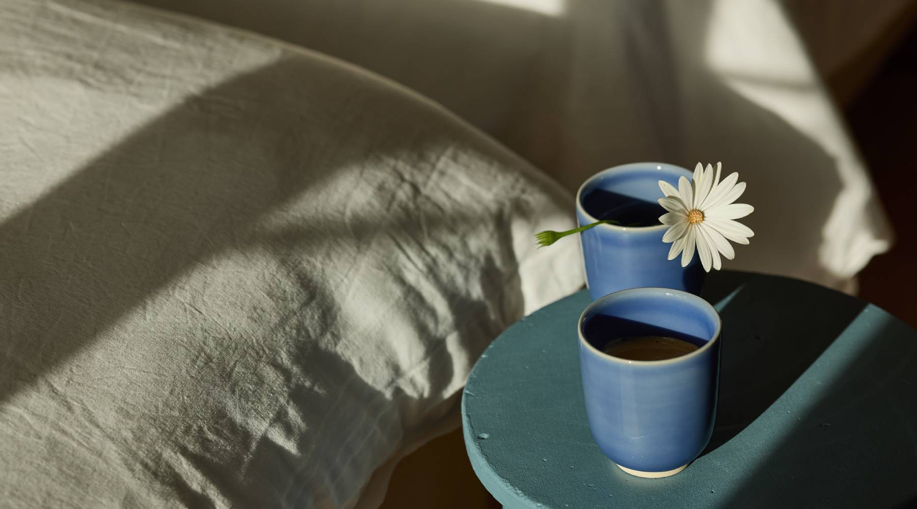 Zwei blaue Keramikbecher mit Kaffee auf einem kleinen petrolfarben gestrichenen Beistelltisch im Sonnenlicht, einer davon mit einer einzelnen weißen Margerite. Im Hintergrund ein helles Leinensofa mit weichen Schattenspielen.