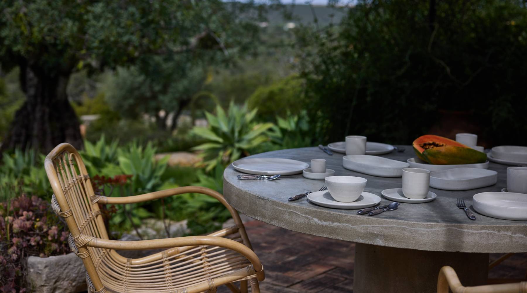 Gedeckter Tisch mit grauem Geschirr und Rattanstuhl im grünen Garten