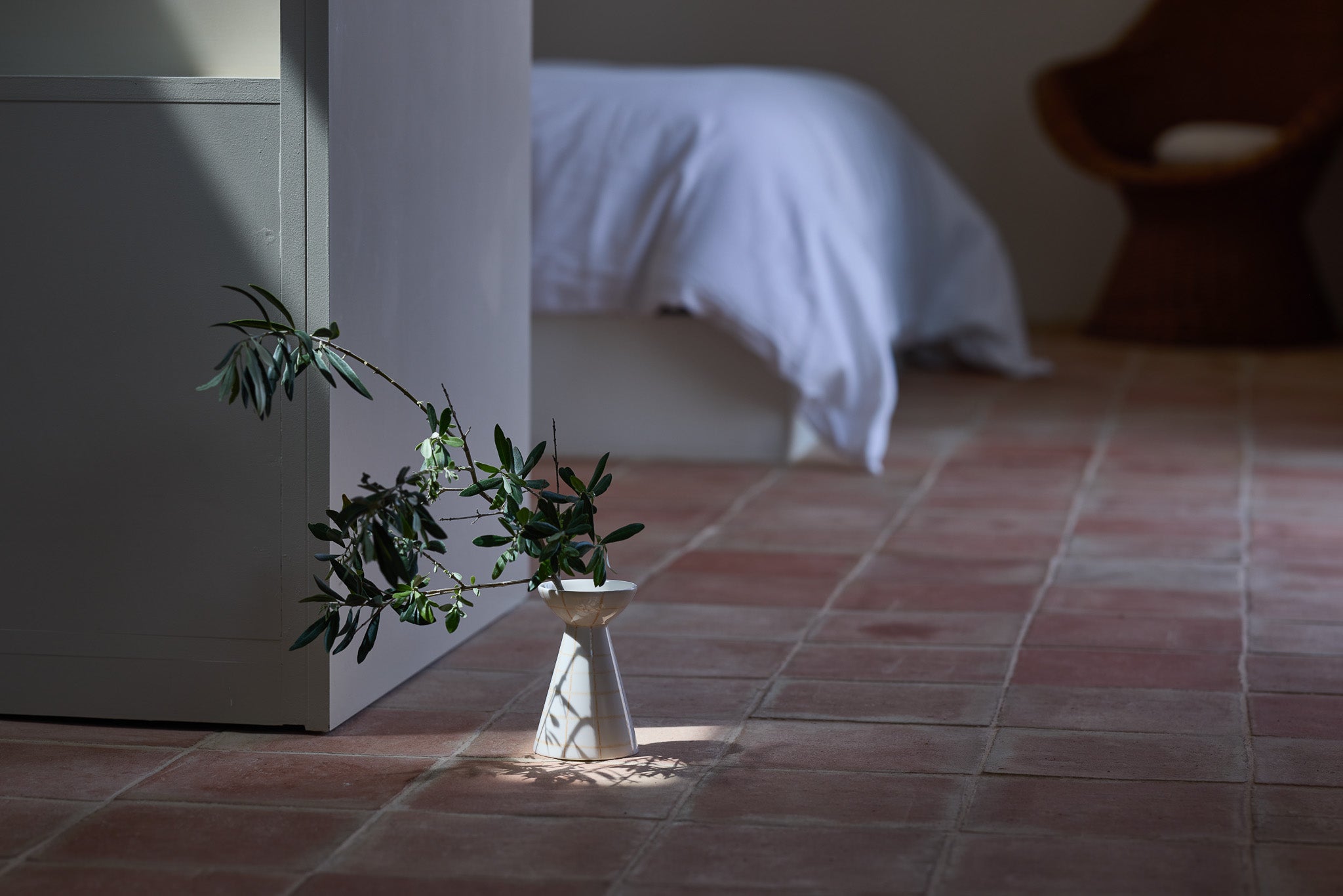 Eine schlanke, taillierte Keramikvase in Weiß mit einem Olivenzweig auf einem rustikalen Terrakottaboden. Ein Lichtstrahl fällt auf die Vase und wirft einen markanten Schatten. Im unscharfen Hintergrund ein Bett mit weißer Bettwäsche und ein Holzstuhl – eine ruhige, mediterrane Schlafzimmeratmosphäre.