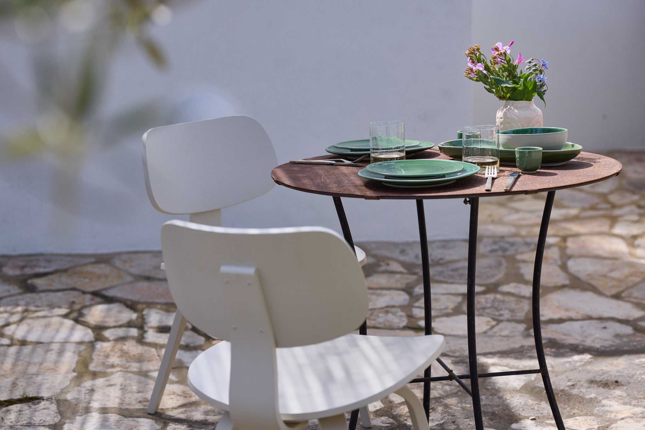 Kleiner runder Bistrotisch mit schwarzem Eisengestell auf einem mediterranen Steinpflaster-Außenbereich. Der Tisch ist für zwei Personen gedeckt mit grün-glasiertem Keramikgeschirr, gerippten Wassergläsern und dunklem Besteck. Eine rosafarbene Keramikvase mit bunten Wildblumen dient als Tischdekoration. Ein weißer moderner Holzstuhl steht vor dem Tisch, im Hintergrund eine weiße Wand im hellen Sonnenlicht.