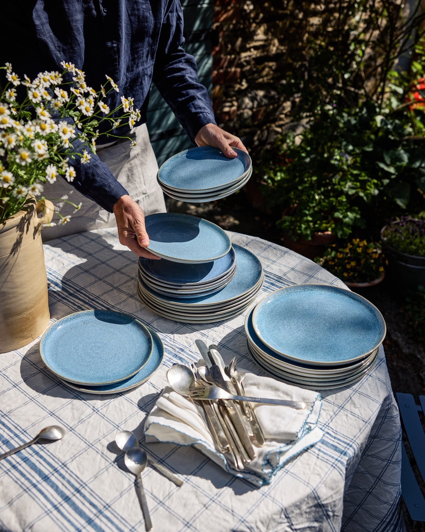 Person deckt einen Gartentisch mit blau-weiß kariertem Leinentischtuch ein und stapelt blaue Keramikteller. Auf dem Tisch liegen weiteres Geschirr, silbernes Besteck und weiße Stoffservietten. Links ein Steinkrug mit Kamillenblüten, im Hintergrund ein bepflanzter Garten.