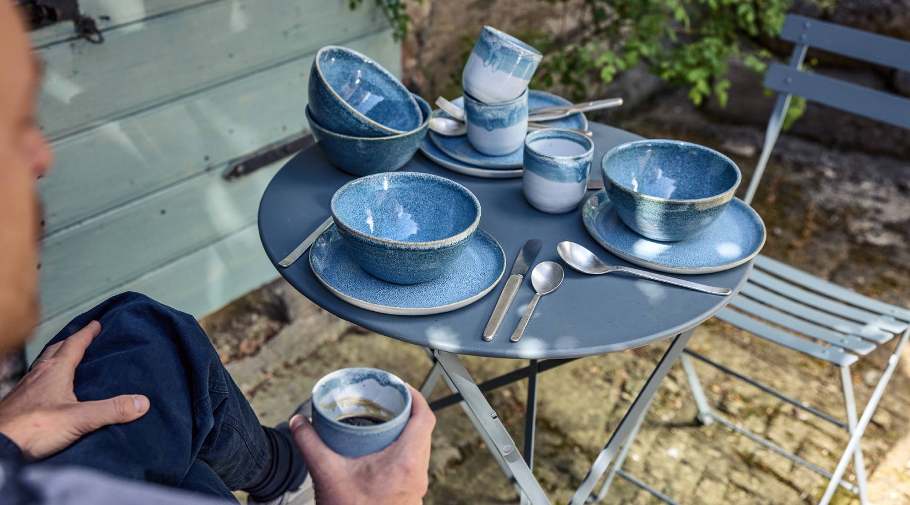 Person hält einen blauen Keramikbecher mit Verlaufsglasur und Kaffee, dahinter ein grauer Metallklapptisch mit blauem Keramikgeschirr (Schalen, Teller und Becher) sowie silbernem Besteck. Rechts ein passender Klappstuhl, im Hintergrund eine mintgrüne Holzwand.