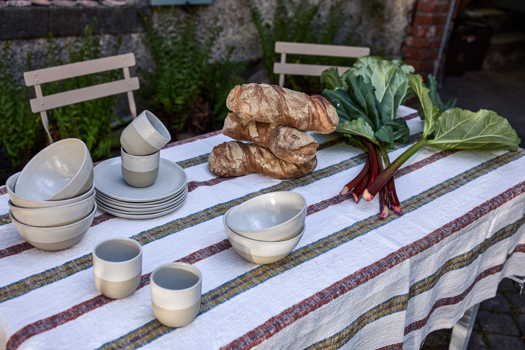 Outdoortisch mit bunt gestreiftem Leinentischtuch, darauf gestapeltes hellgraues Keramikgeschirr (Schalen, Teller und Becher), rustikale Croissants sowie ein frischer Rhabarberstrauß mit großen Blättern. Im Hintergrund zwei beige Gartenstühle vor einer Natursteinmauer mit Farnbewuchs.