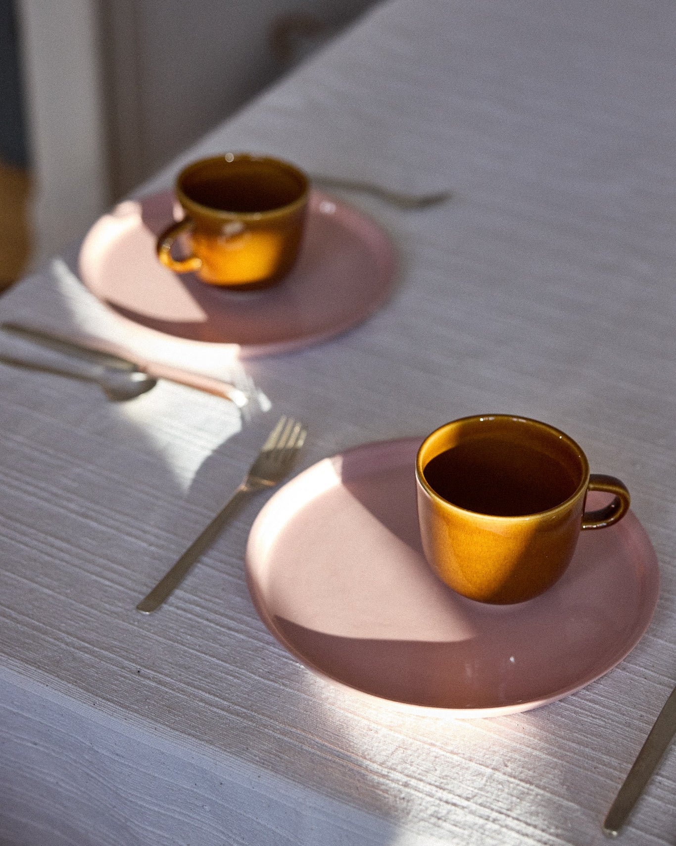 Zwei bernsteinfarbene Keramiktassen mit Kaffee auf rosafarbenen Keramiktellern, auf einem hellgrauen Holztisch im Sonnenlicht. Daneben eine weiße Leinenserviette mit silbernem Besteck.