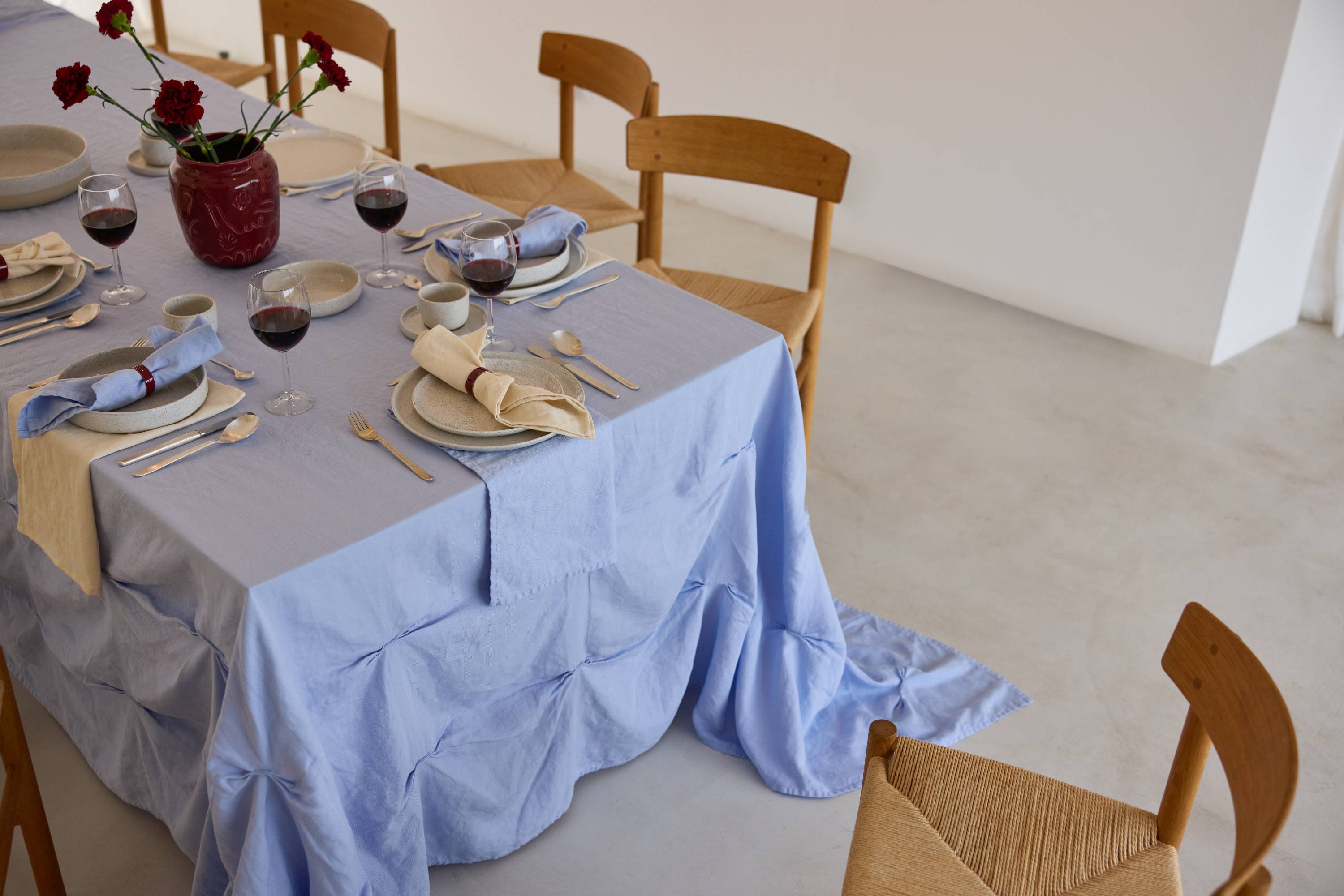 Gedeckter Esstisch mit hellblauer Leinentischdecke, Keramikgeschirr, Holzbesteck, Rotweingläsern und roter Blumenvase, umgeben von Holzstühlen