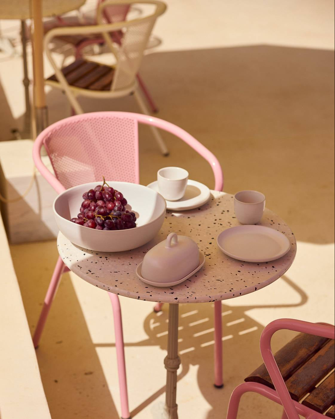 Runder Bistrotisch aus Terrazzo mit pinken Metallbeinen, auf dem ein weißes Geschirr-Set arrangiert ist: eine Schale mit roten Weintrauben, eine Espressotasse auf Unterteller, ein mattrosafarbener Becher sowie ein Butterdöschen und ein kleiner Teller in gedämpftem Rosa. Dahinter ein Armlehnstuhl mit pinkfarbenem Metallgestell und gelochter Rückenlehne. Die Szene wirkt wie eine sommerliche Terrassensituation in warmem, sandfarbenem Umgebungslicht.